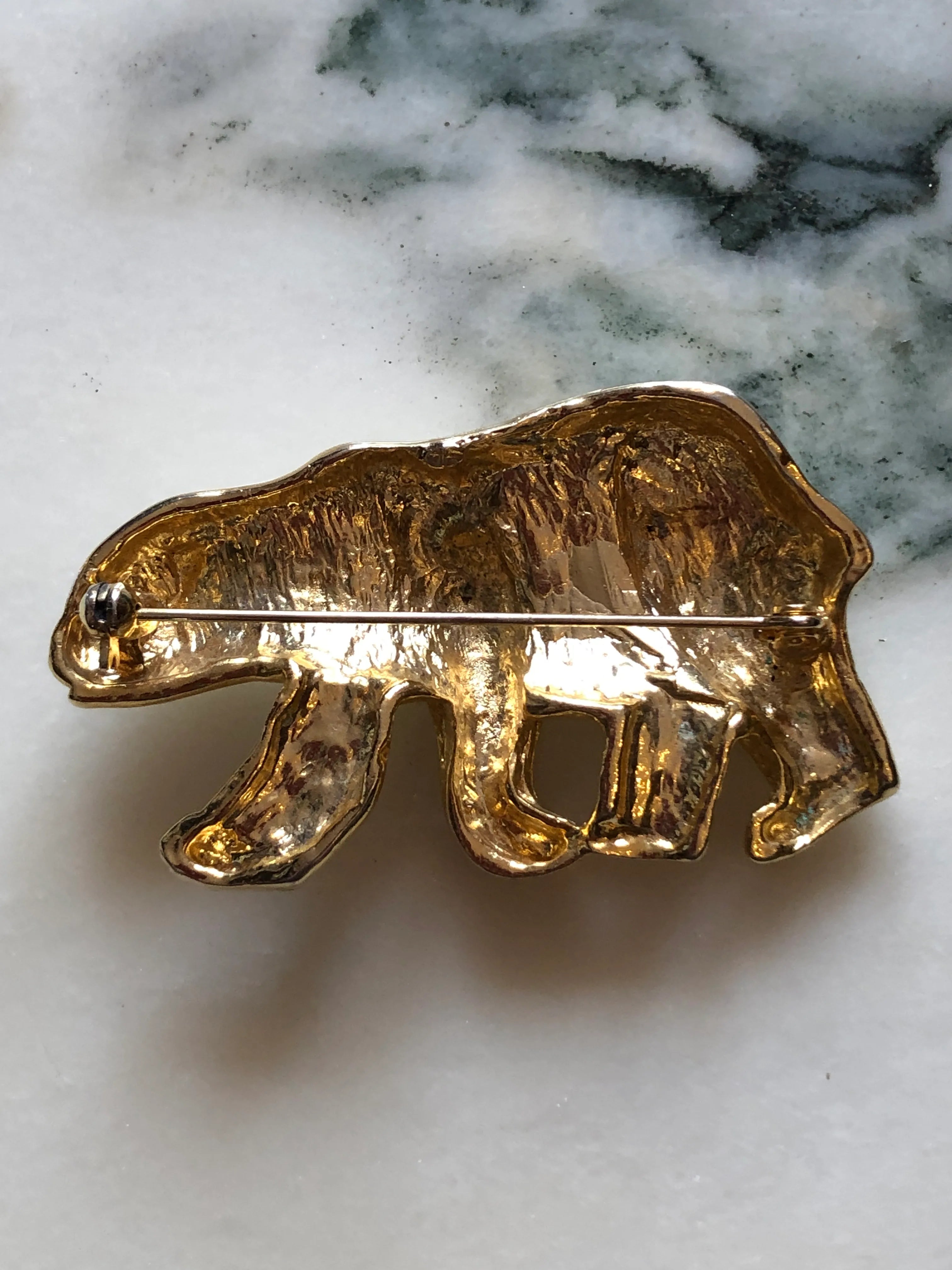 The back view of Broochella Orso Bianco vintage white enamel polar bear brooch with gold-tone details and a green crystal eye. The background is white marble