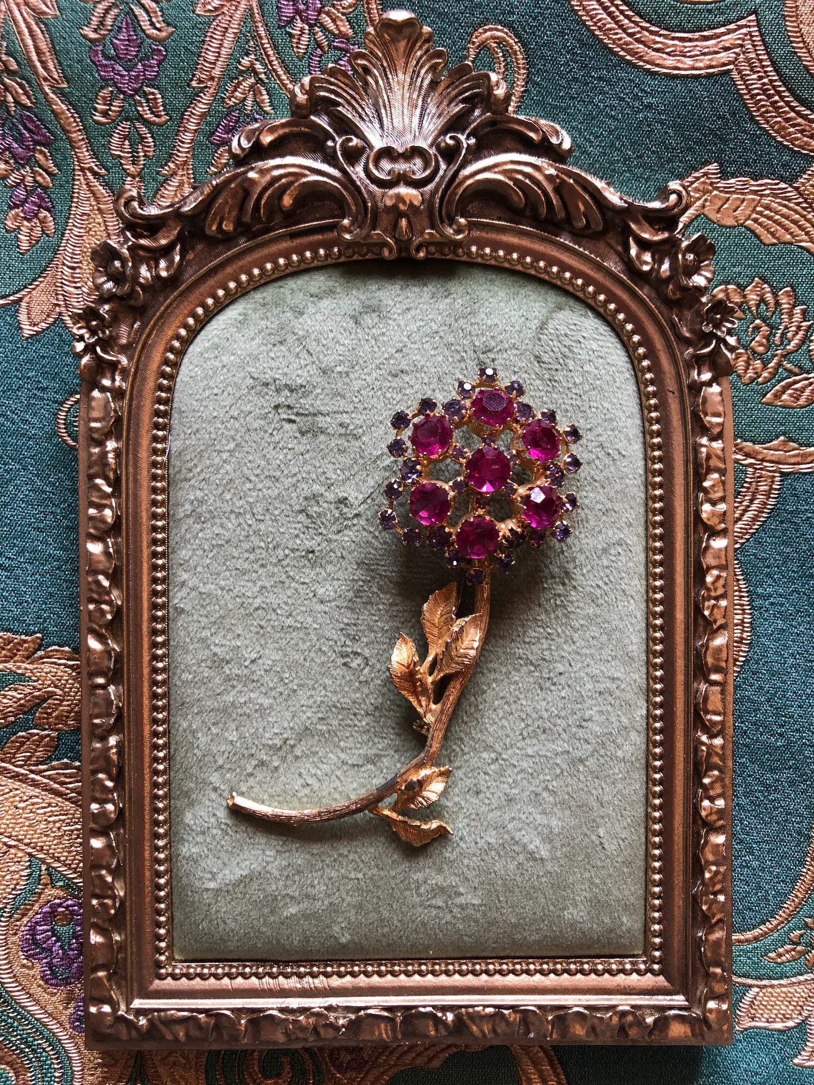 Broochella Ortensia Reale vintage gold-tone flower brooch with a long stem, textured leaves and fuchsia and purple crystals. Displayed inside an ornate antique bronze frame against a patterned teal fabric background.