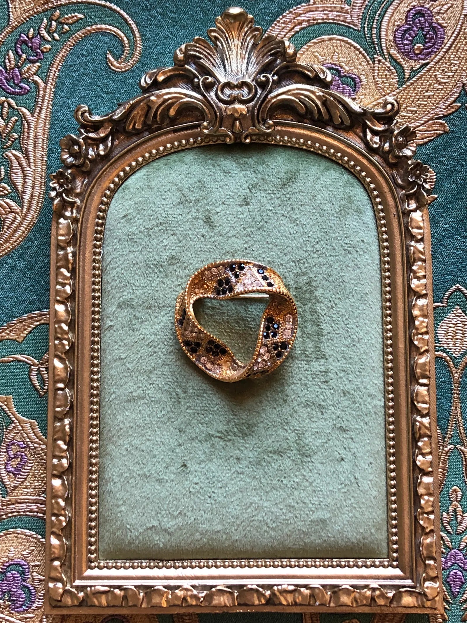 Broochella Ottavia Vintage Black & Crystal Swirl Brooch featuring black and clear crystals set into a sculptural gold-tone circular design. Pictured on a green velvet tray with an ornate gold frame. The background is green and gold damask wallpaper. 