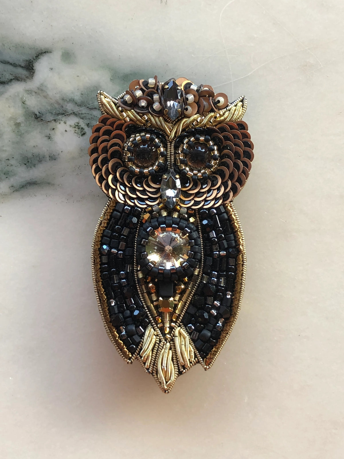 Broochella Owl Brooch Pin hand-beaded in black, bronze and gold with sequins, crystals and intricate beadwork. Pictured on a white marble surface.