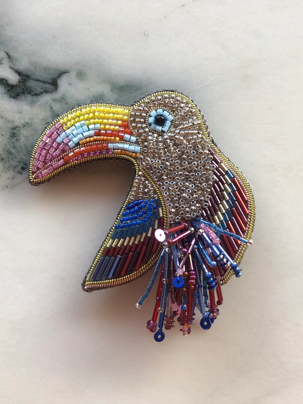Broochella Pappagallo Parrot Brooch, hand-beaded with metallic gold, pink, blue, yellow and red beads, featuring a colourful curved beak and beaded tail detail.