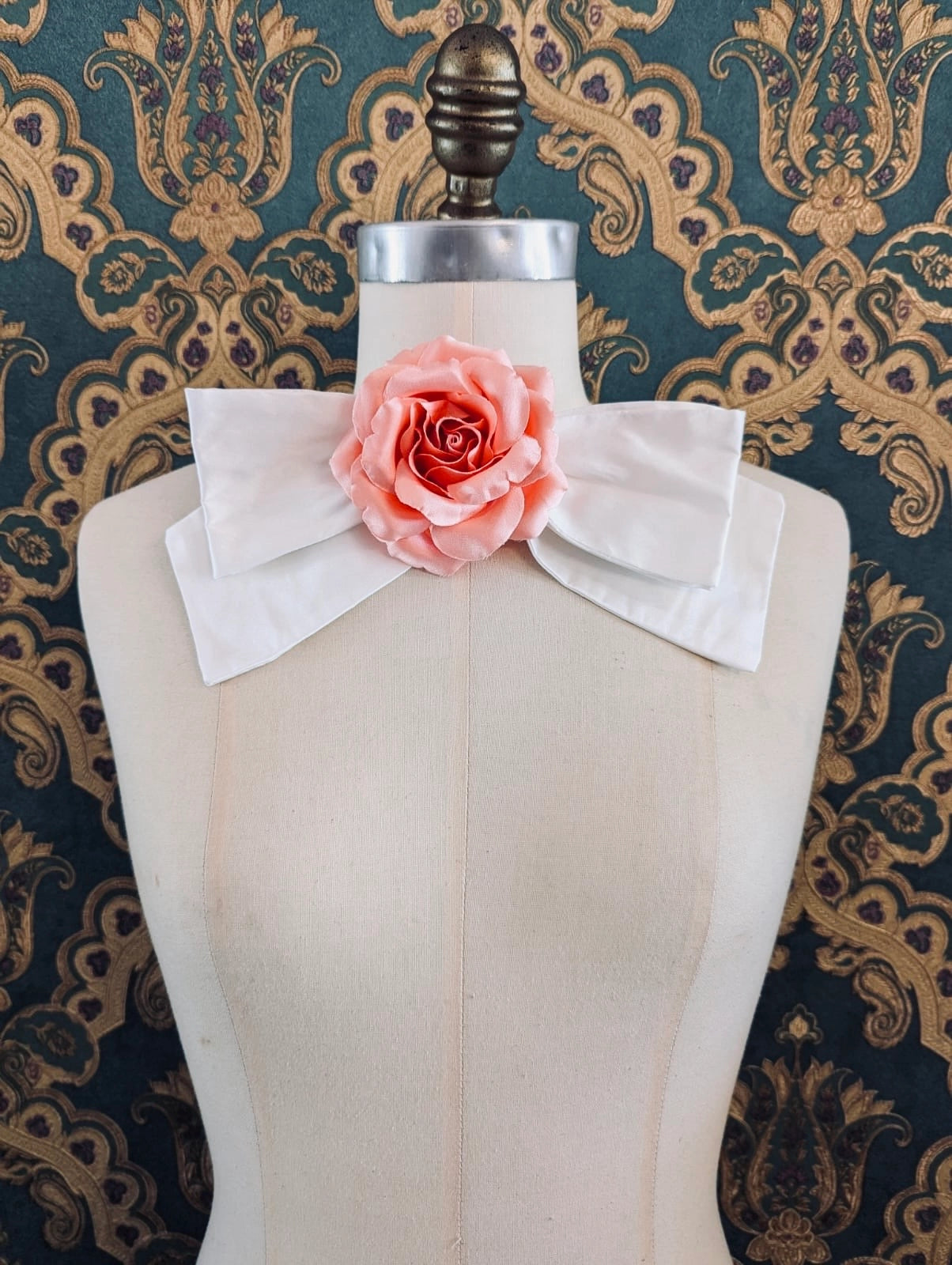 Broochella peaches and cream handmade ivory silk bow brooch, with peach flower embellishment.  The background is green and gold damask wallpaper.