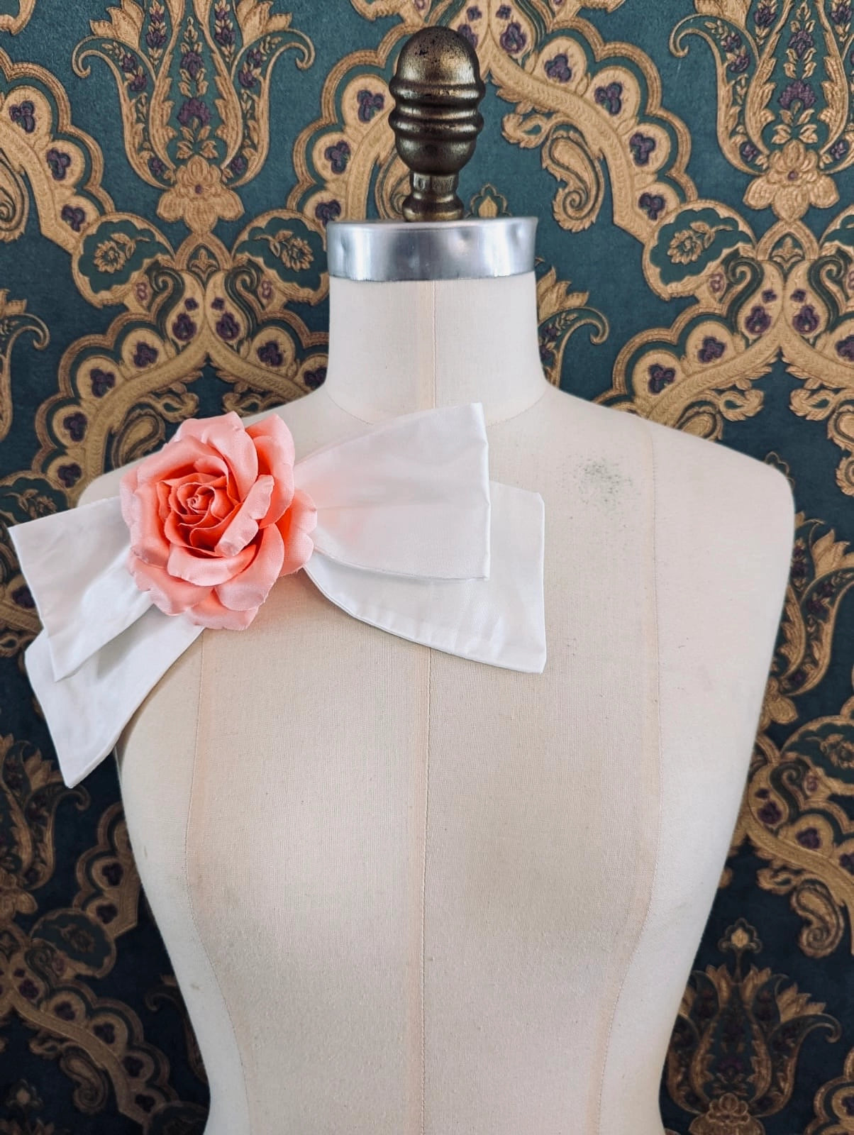 Broochella peaches and cream handmade ivory silk bow brooch, with peach flower embellishment.  The background is green and gold damask wallpaper.