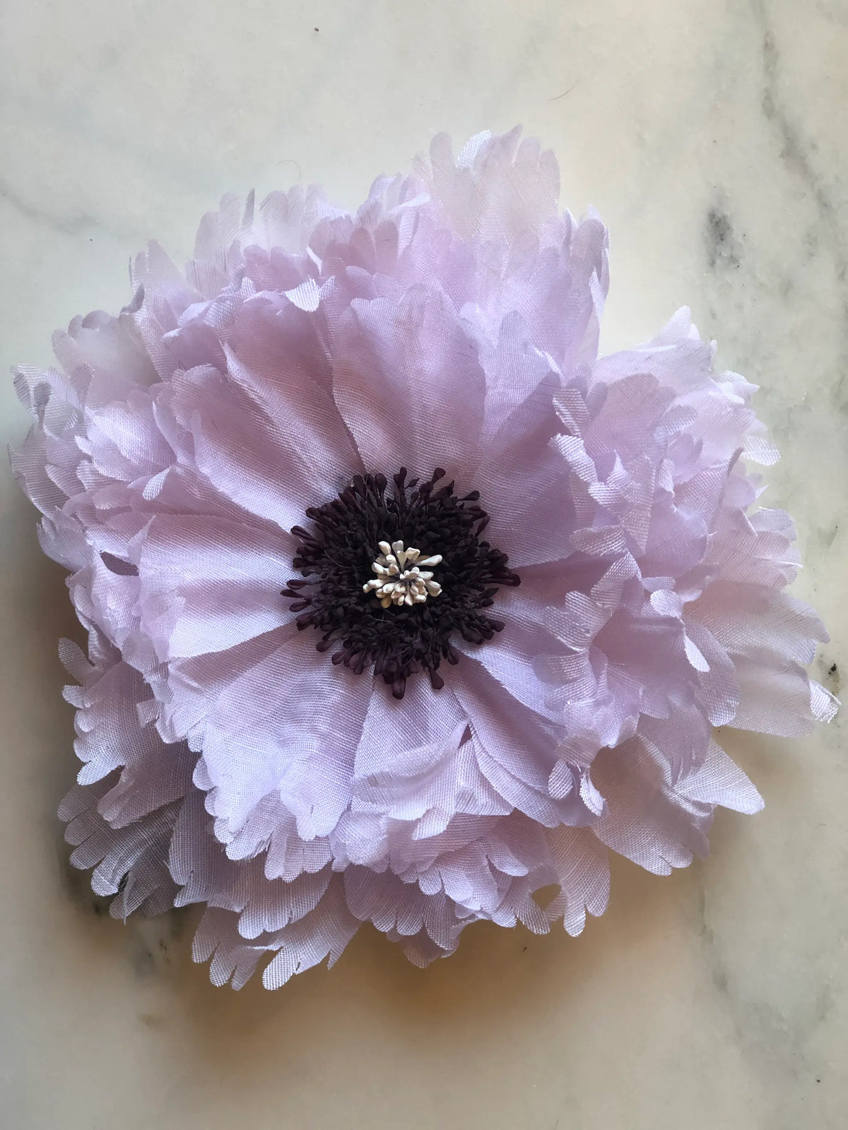 Broochella Peone Large Flower Brooch in soft blush pink, featuring oversized layered fabric petals with ruffled edges, a dark plum stamen centre and delicate cream accents on a marble background.