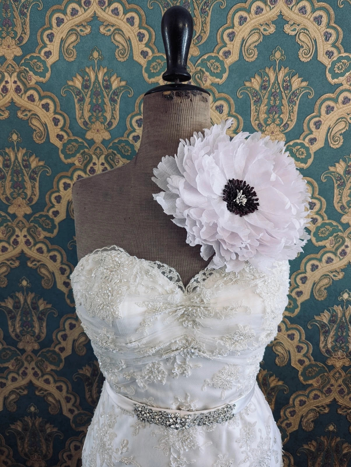 Broochella bridal peone large pink handmade flower brooch. Pictured on a mannequin that is wearing a white, beaded strapless wedding dress. The background is green and gold damask wallpaper. 