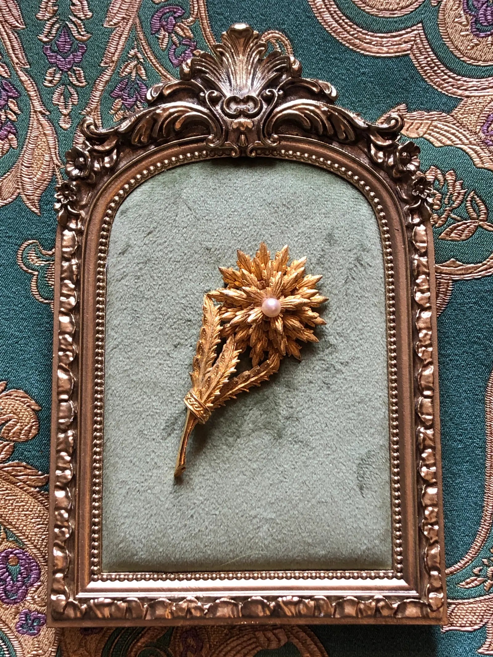 A vintage Broochella Perla d'oro gold leaf and acanthus leaf brooch with a central pink pearl, displayed inside an ornate antique bronze picture frame against a teal patterned fabric background.