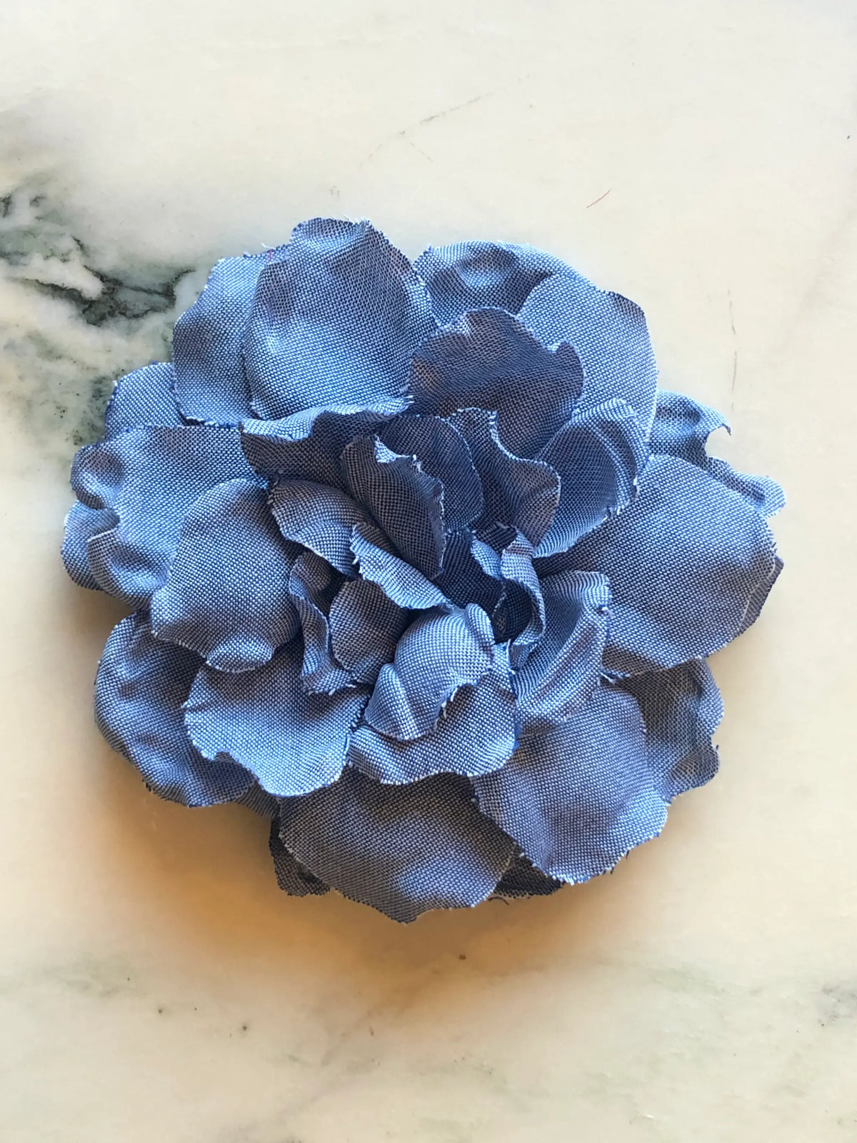 Broochella Pervinca Denim Flower Brooch in mid blue denim, featuring full layered sculptural petals with softly curved edges on a cream marble background.