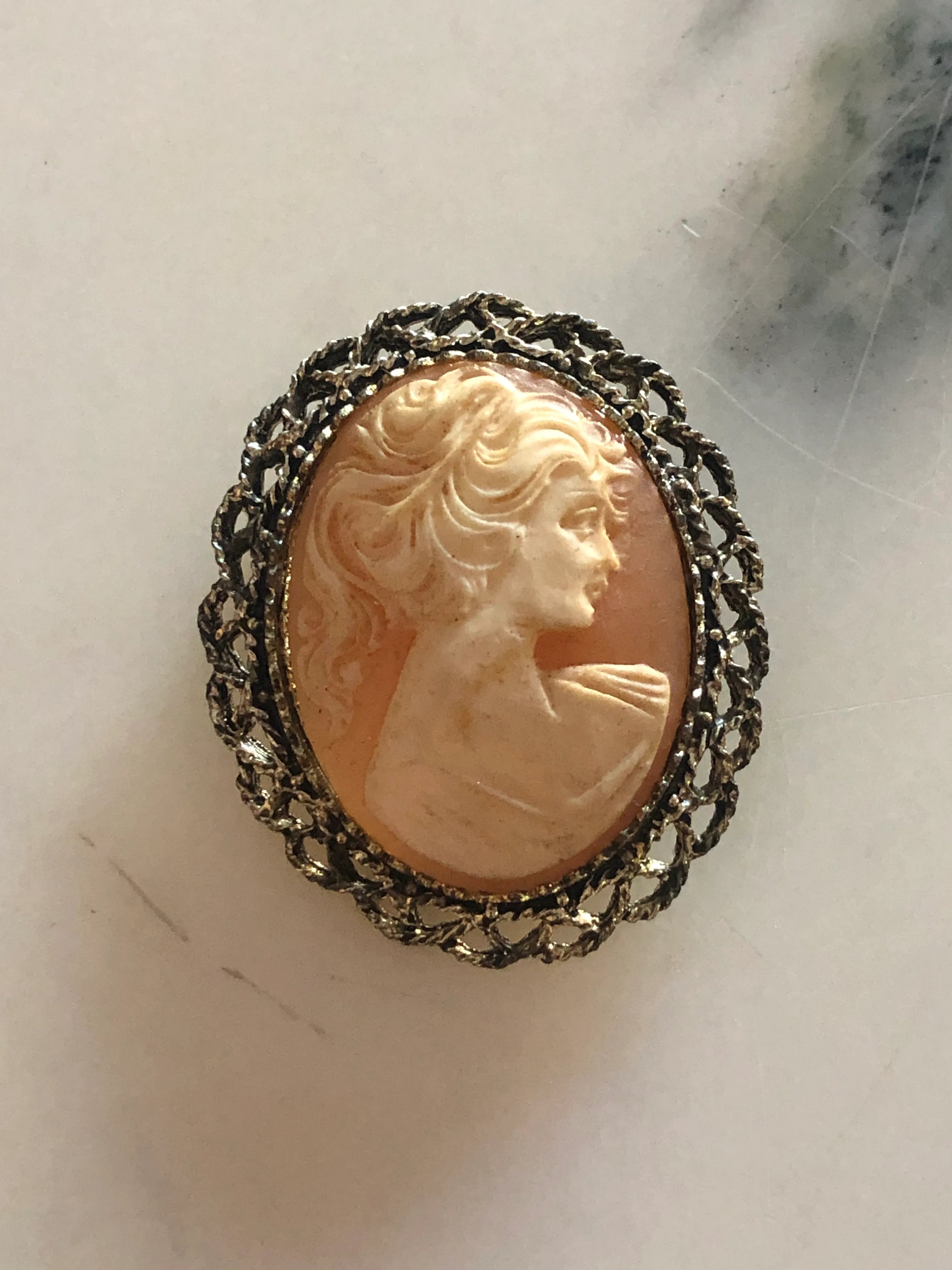 A Broochella Pesca oval vintage cameo brooch featuring a carved profile of a woman on a peach-colored cabochon, surrounded by a dark oxidized silver filigree border, shown on a light marble surface.