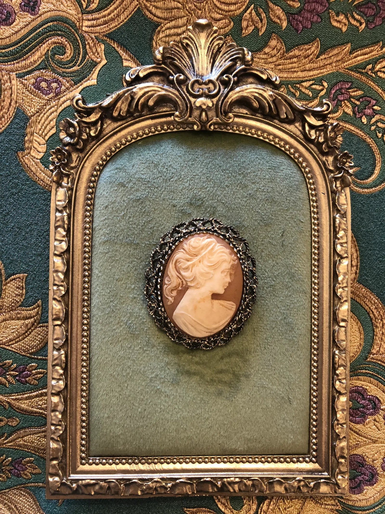 Broochella Pesca vintage cameo brooch in coral and ivory. It has an ornate gunmetal frame. The brooch is pinned to a green velvet tray, with a gold frame.  The background is green nd gold damask wallpaper. 
