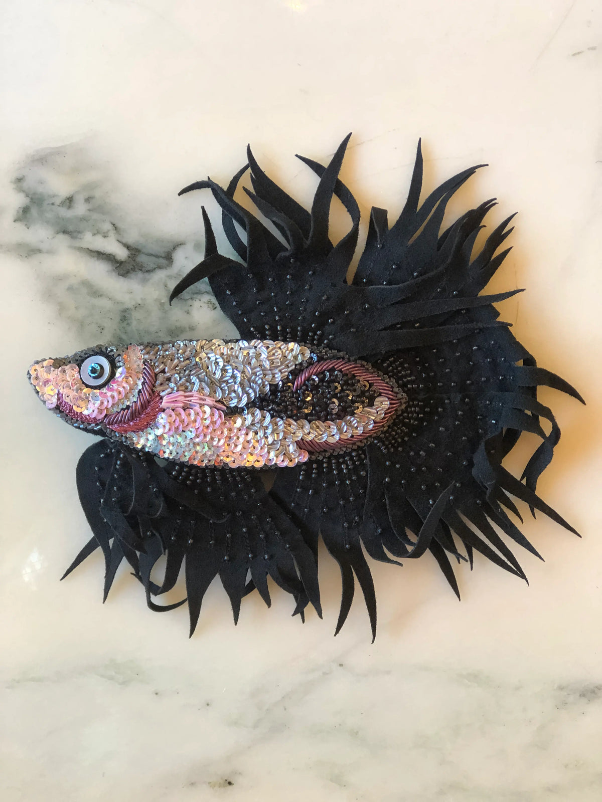 Broochella Pesce Siamese Fish Brooch featuring an ornate Siamese fighting fish with hand-beaded black fins, sculptural fringe and a shimmering pink, silver and black sequin body on a cream marble background.