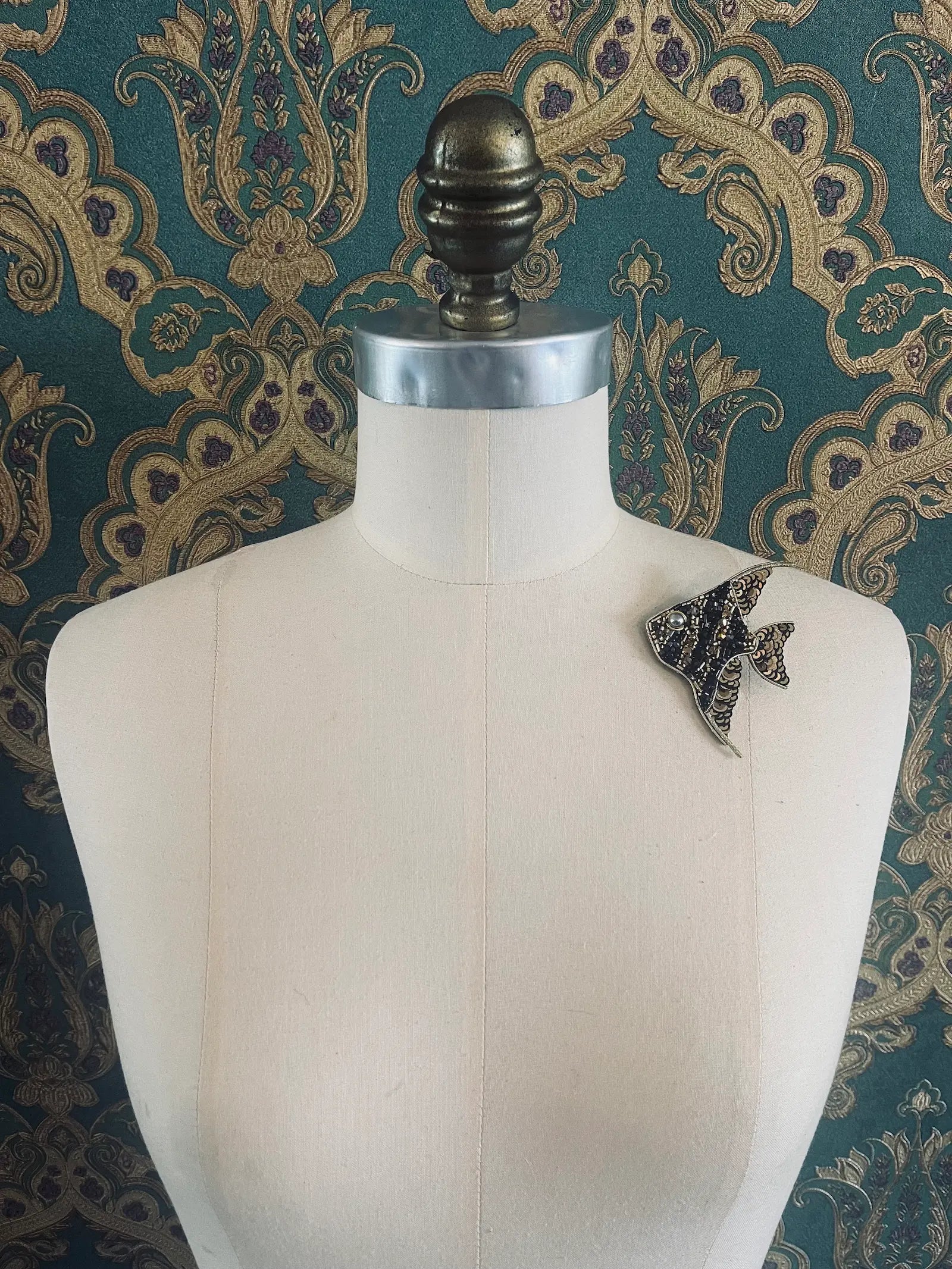 Broochella Pesciolino hand beaded gold and black designer brooch. Pictured on a mannequin. The background is green and gold damask wallpaper