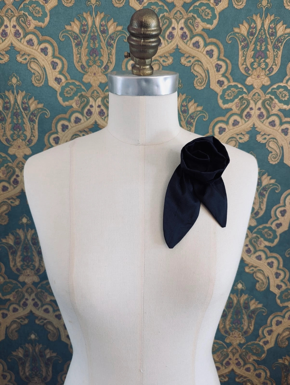 A Broochella Petalo black, small, handmade silk  rose-shaped flower brooch with petals. It is displayed on a cream-coloured fabric dress form against a dark teal and gold damask patterned wallpaper background.