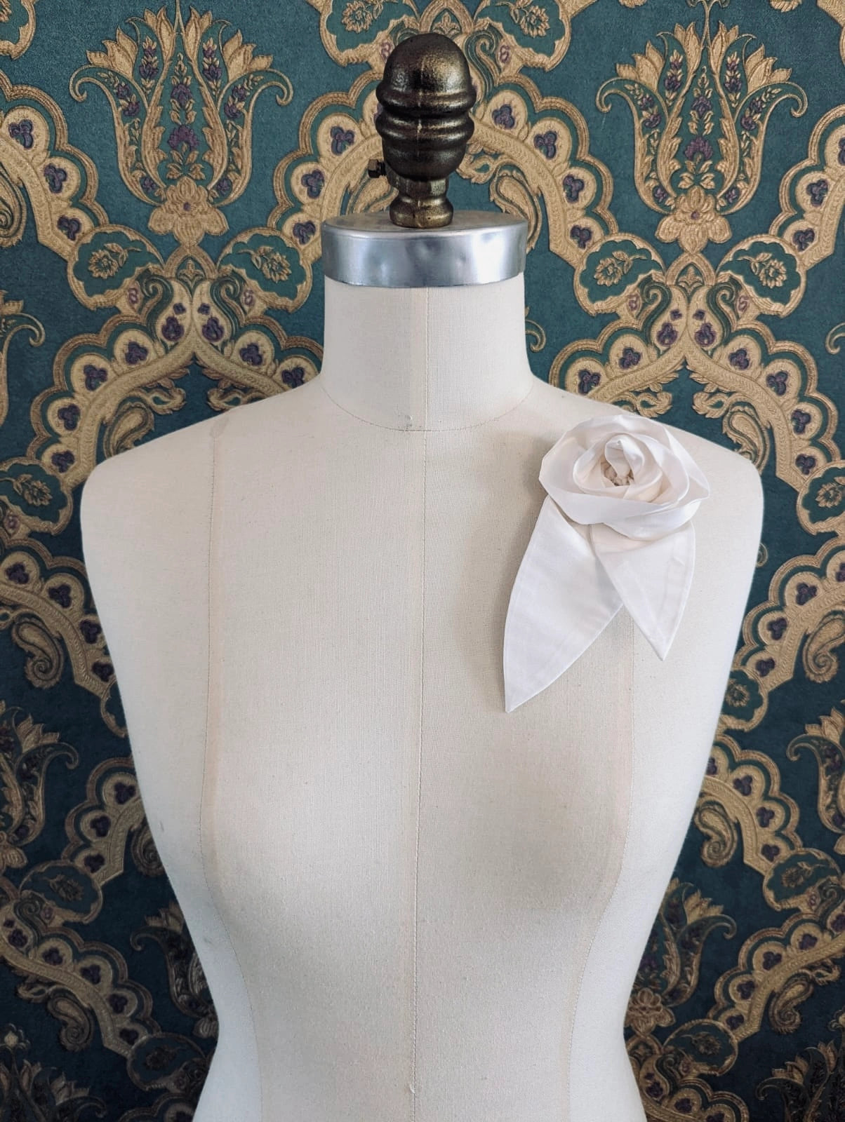 A Broochella Petalo ivory, small, handmade silk  rose-shaped flower brooch with petals. It is displayed on a cream-coloured fabric dress form against a dark teal and gold damask patterned wallpaper background.