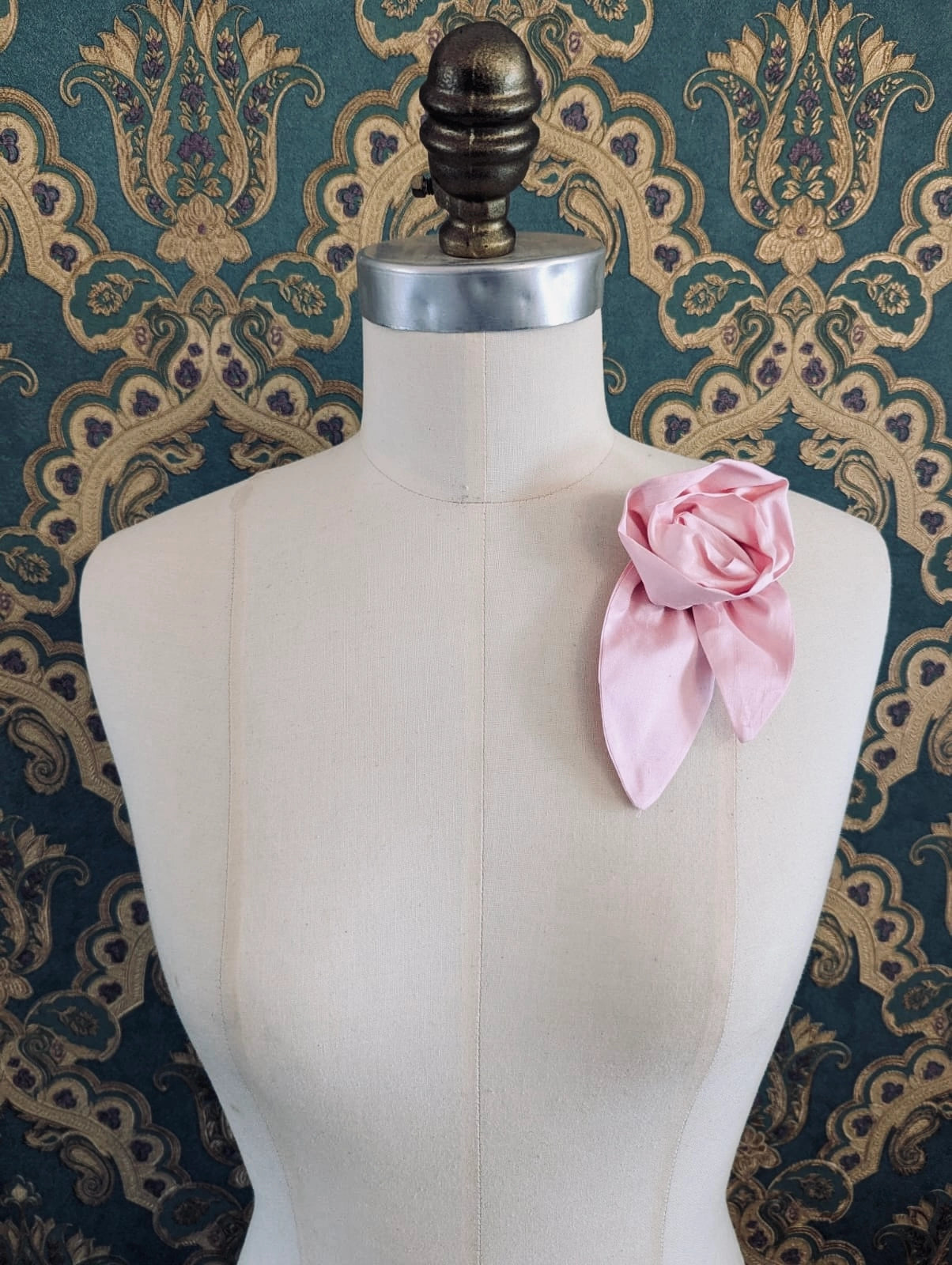 A Broochella Petalo pink, small, handmade silk  rose-shaped flower brooch with petals. It is displayed on a cream-coloured fabric dress form against a dark teal and gold damask patterned wallpaper background.