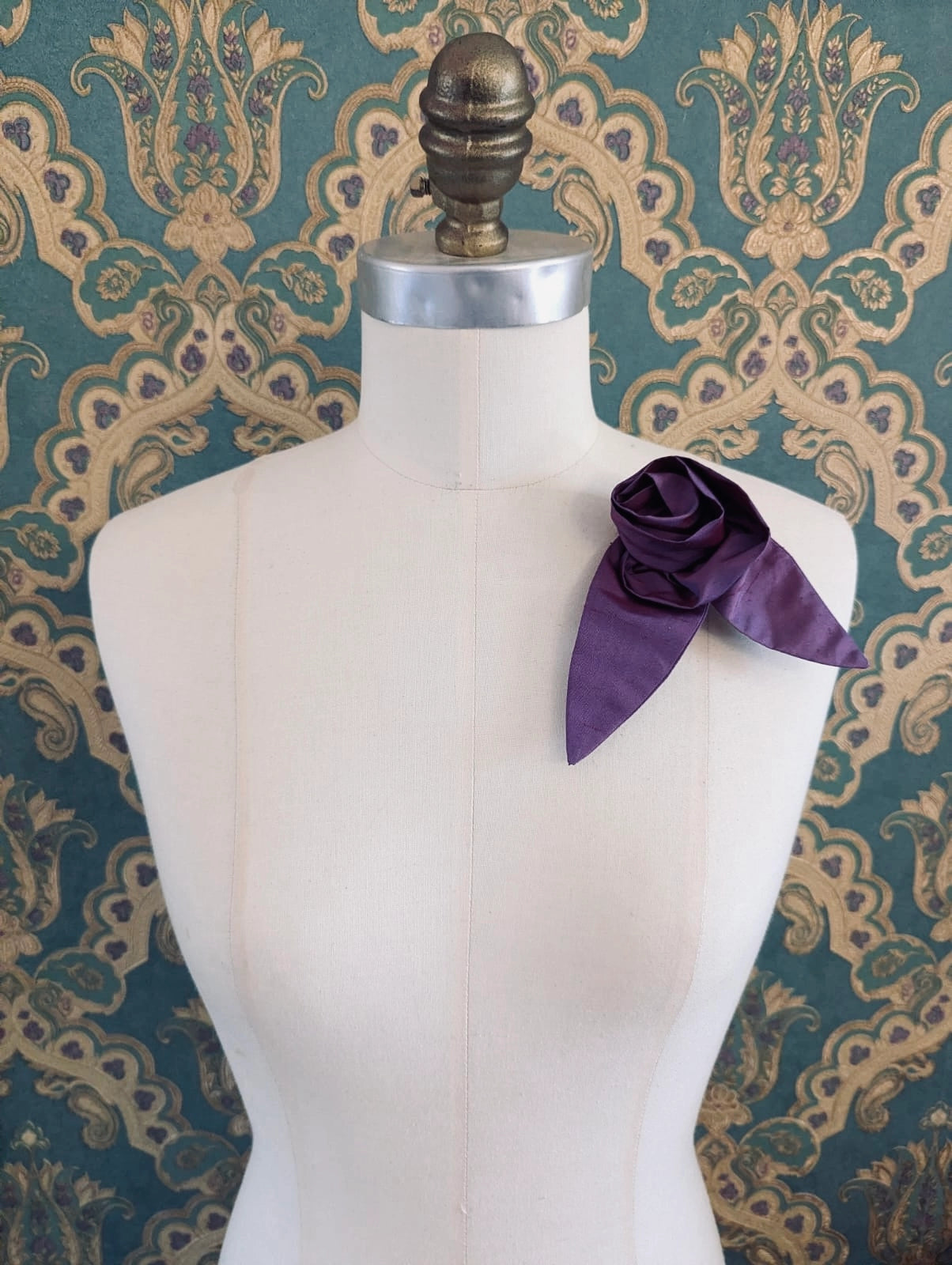 A Broochella Petalo purple, small, handmade silk  rose-shaped flower brooch with petals. It is displayed on a cream-coloured fabric dress form against a dark teal and gold damask patterned wallpaper background.