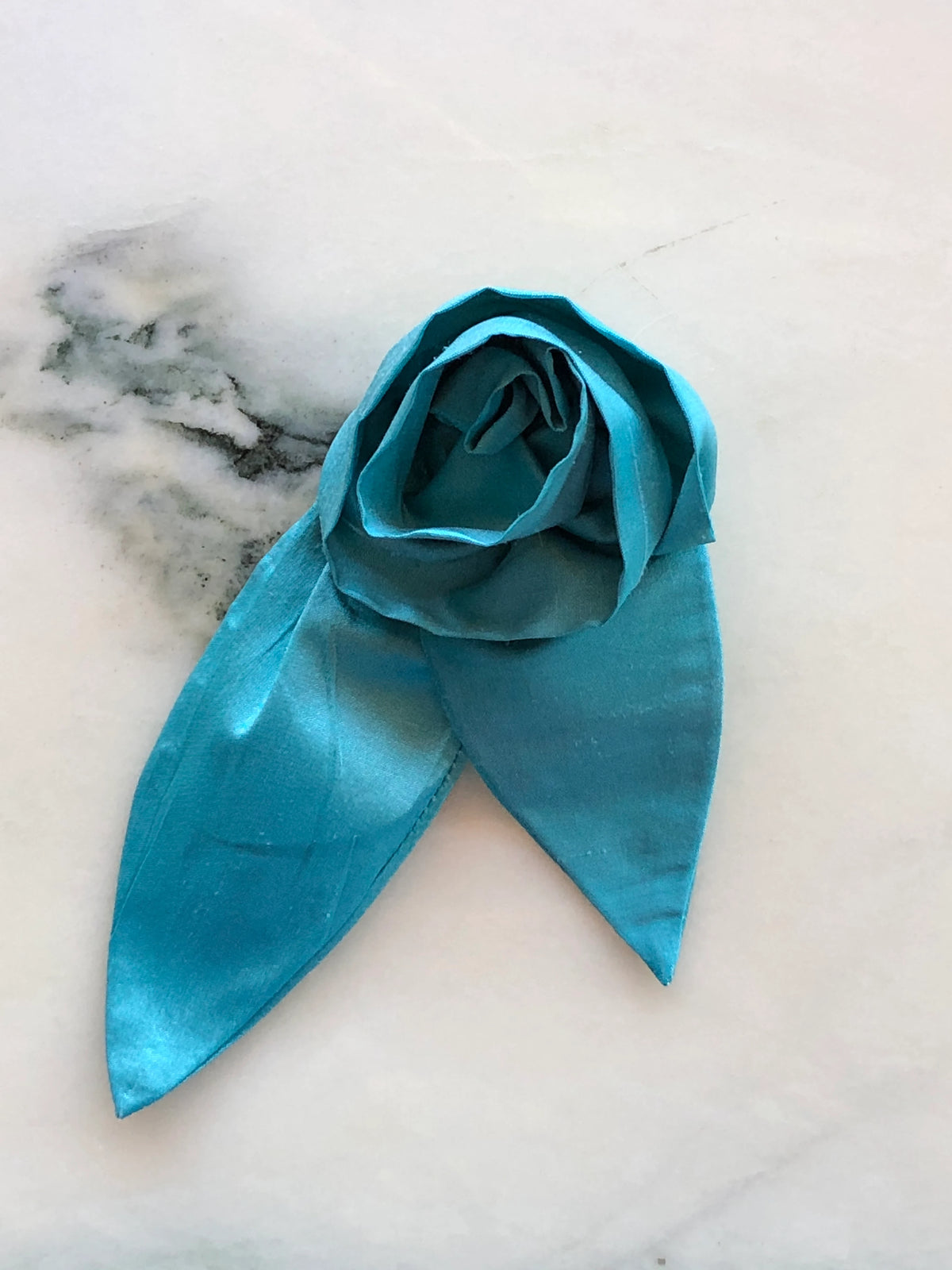 Broochella Petalo Men blue silk flower brooch for men with sculptural rose petals and two pointed silk leaves, photographed on white marble with grey-green veining.
