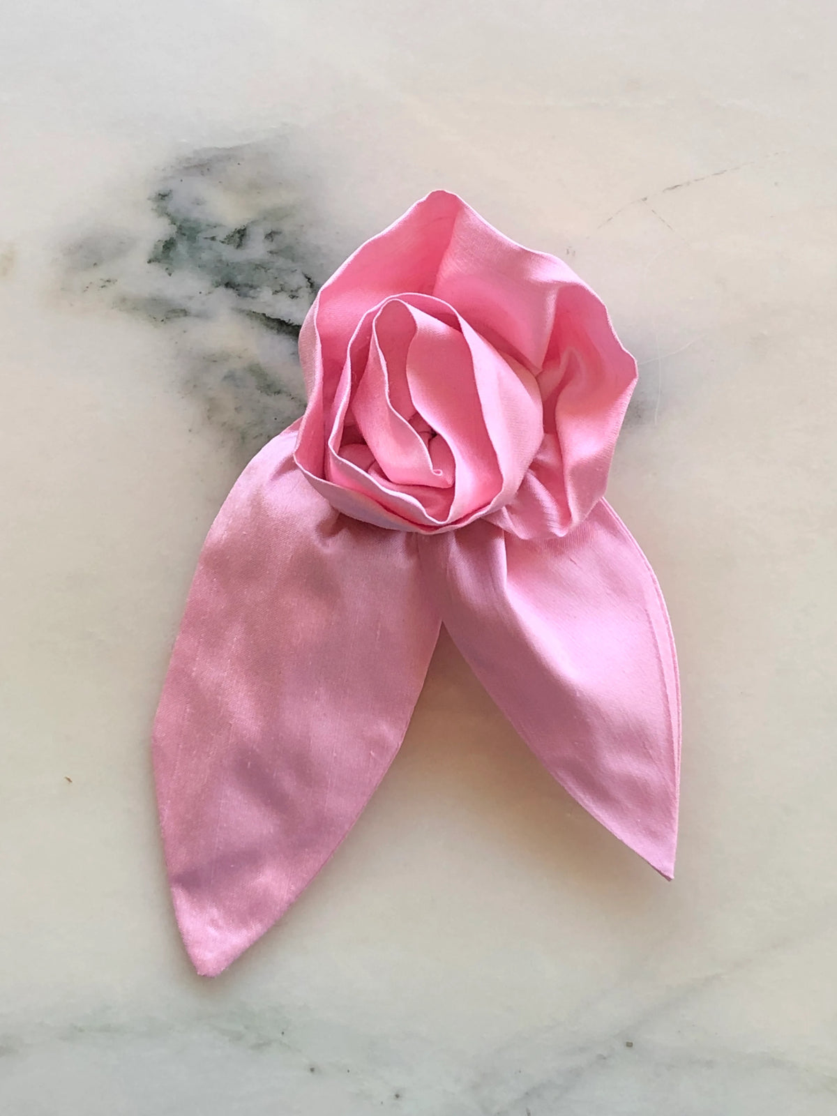 Broochella Petalo Small pink silk flower brooch with sculptural rose petals and two pointed silk leaves, photographed on white marble with grey-green veining.