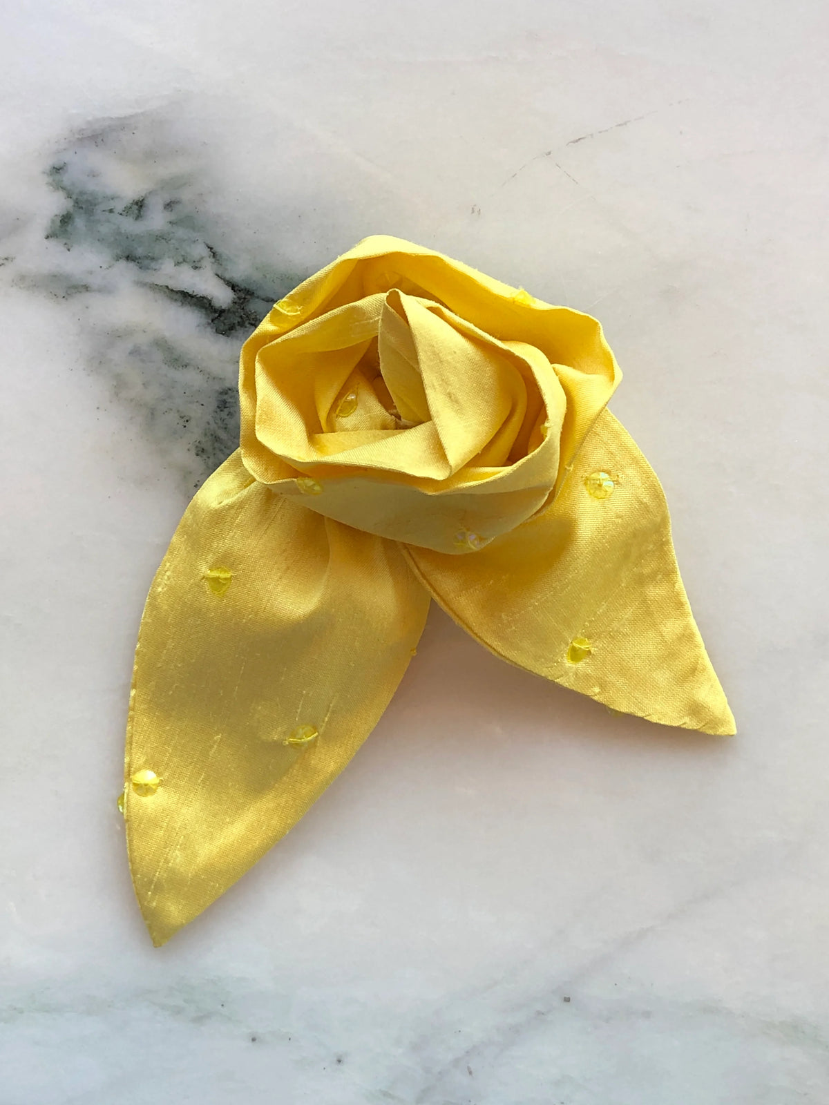 Broochella Petalo Small yellow silk flower brooch with sculptural rose petals, two pointed silk leaves and subtle embroidered details, photographed on white marble with grey-green veining.