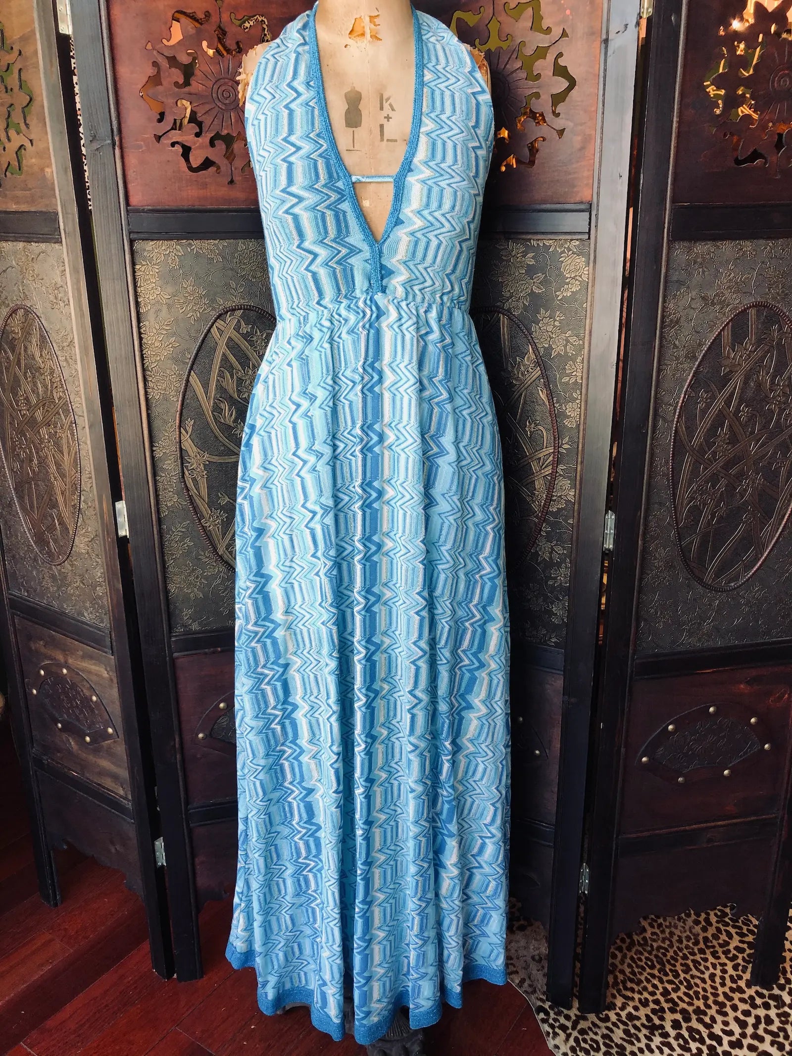 Broochella Pho Firenze maxi dress. Size 
medium. It is a blue, white and silver zigzag pattern and is made from  46% Cotton, 46% Polyester, 8% Lurex. It has a 
halter neckline, is maxi length and has a lightweight, flowing silhouette. It is pictured on a vintage mannequin The background is an ornate, dark wood, vintage room divider. 