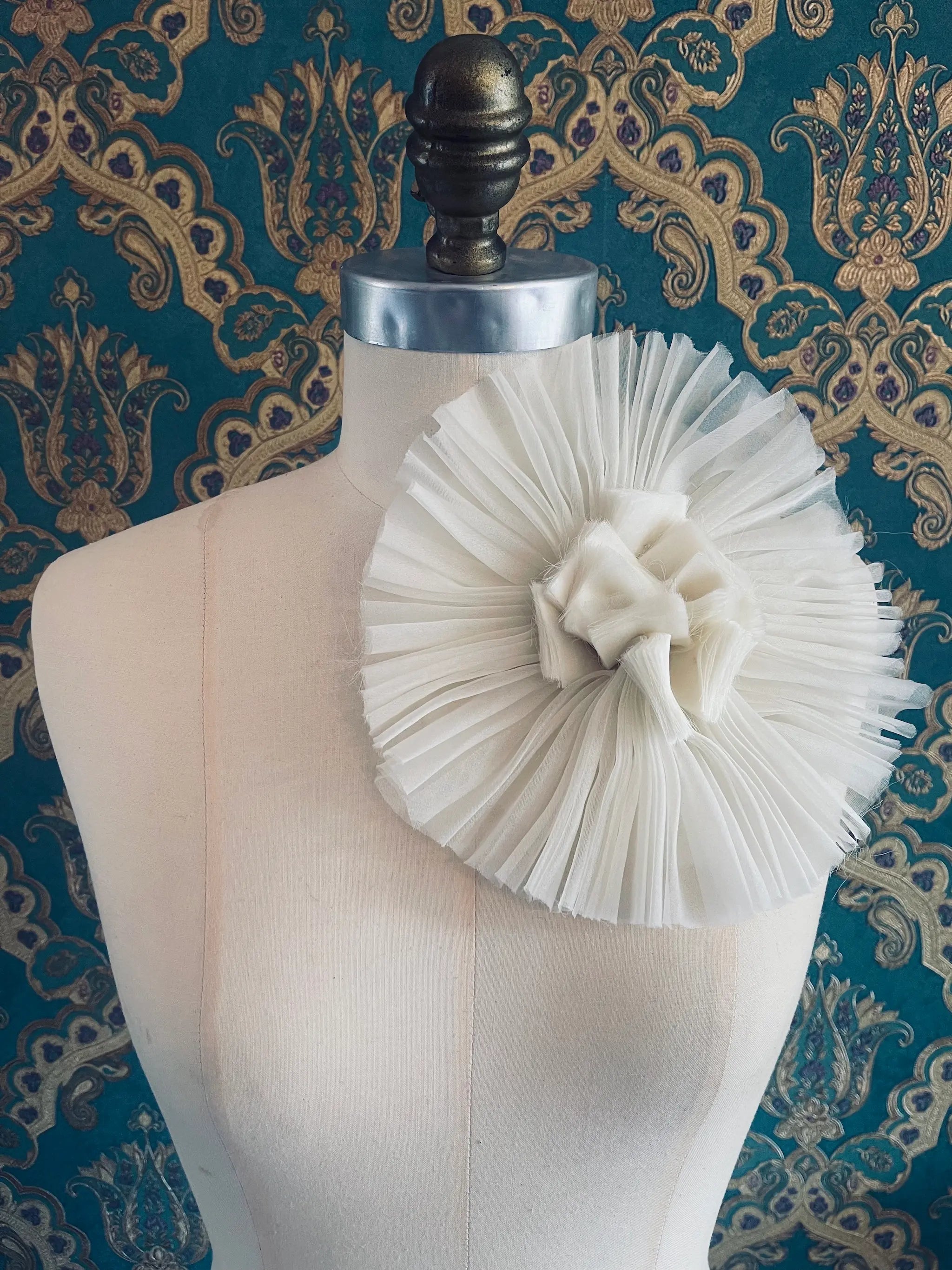 Broochella Pieghettato large handmade fabric flower brooch in white. Styled on a mannequin. The background is green and gold damask wallpaper