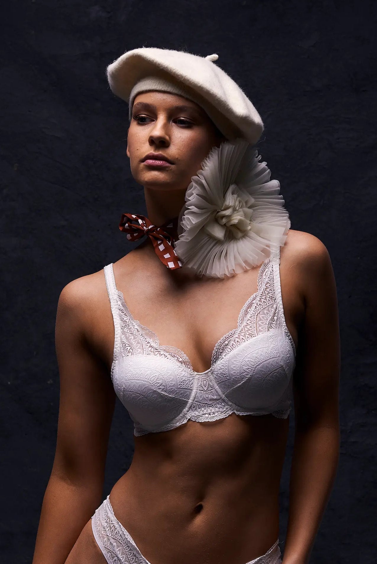 A woman modeling white lace lingerie with a white beret and a large Broochella pieghettato lavender  flower brooch. She is posed against a dark textured studio background.