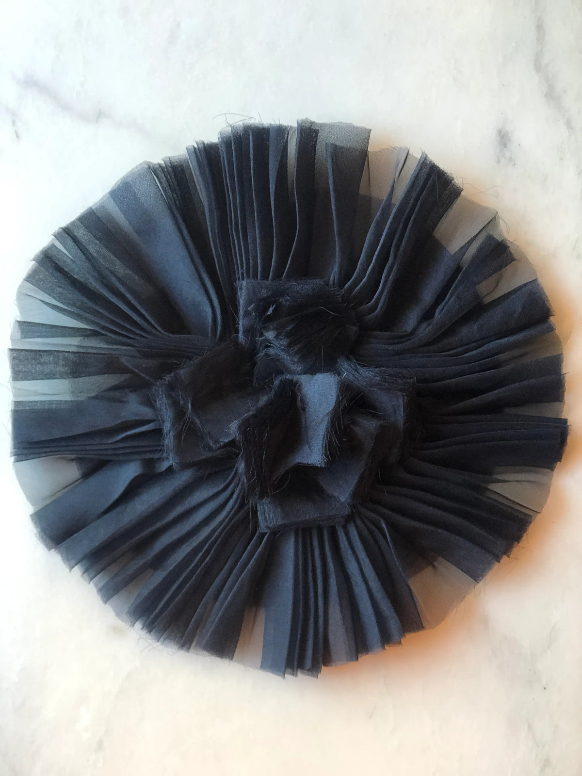 Broochella Men's Pieghettato Large Organza Flower Brooch Pin in black, featuring dramatic pleated organza petals arranged in a circular design with a softly frayed sculptural centre on a marble background