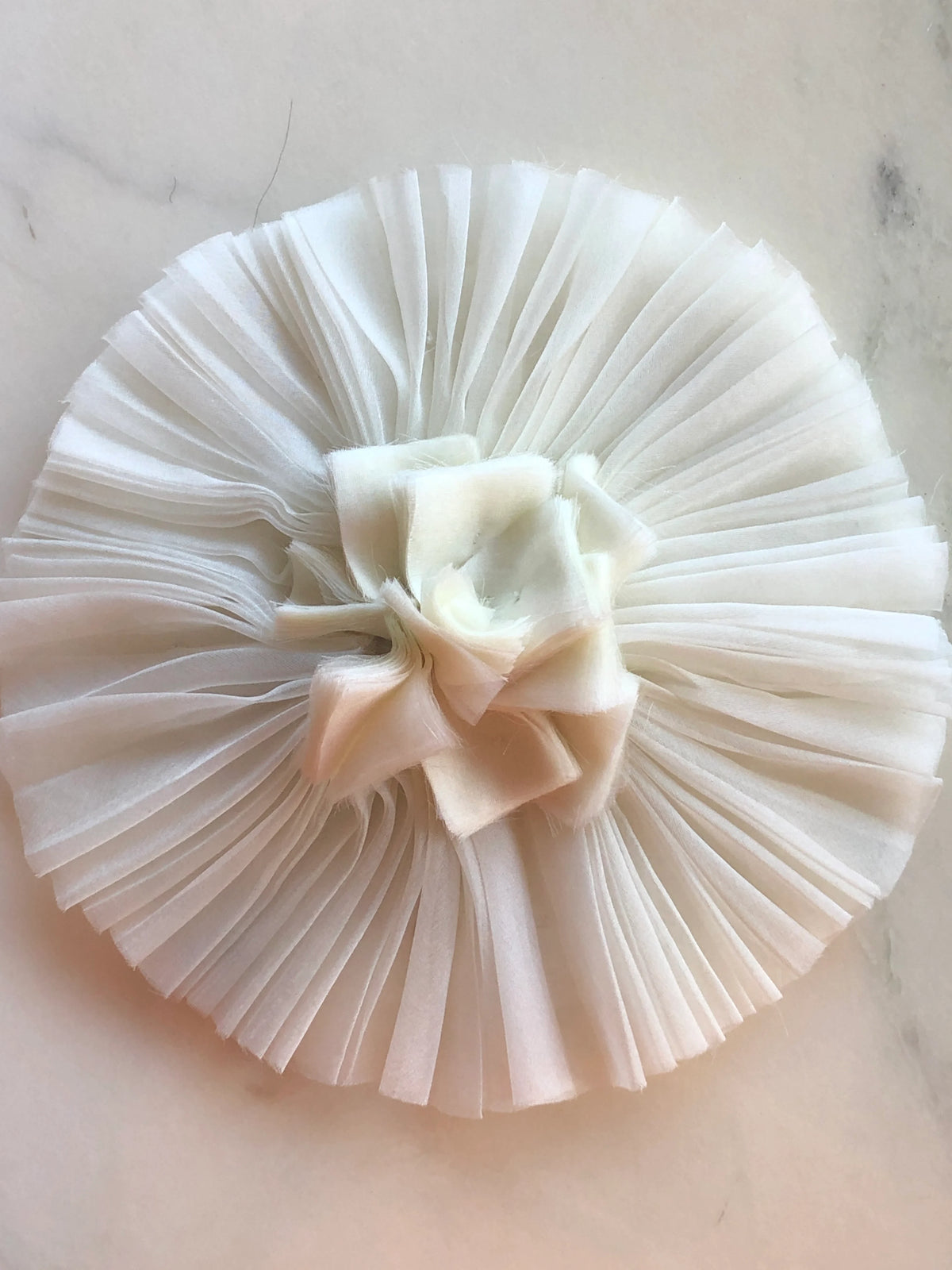Broochella Men's Pieghettato Large Organza Flower Brooch Pin in ivory, featuring pleated organza petals arranged in a circular design with a softly frayed sculptural centre on a marble background.