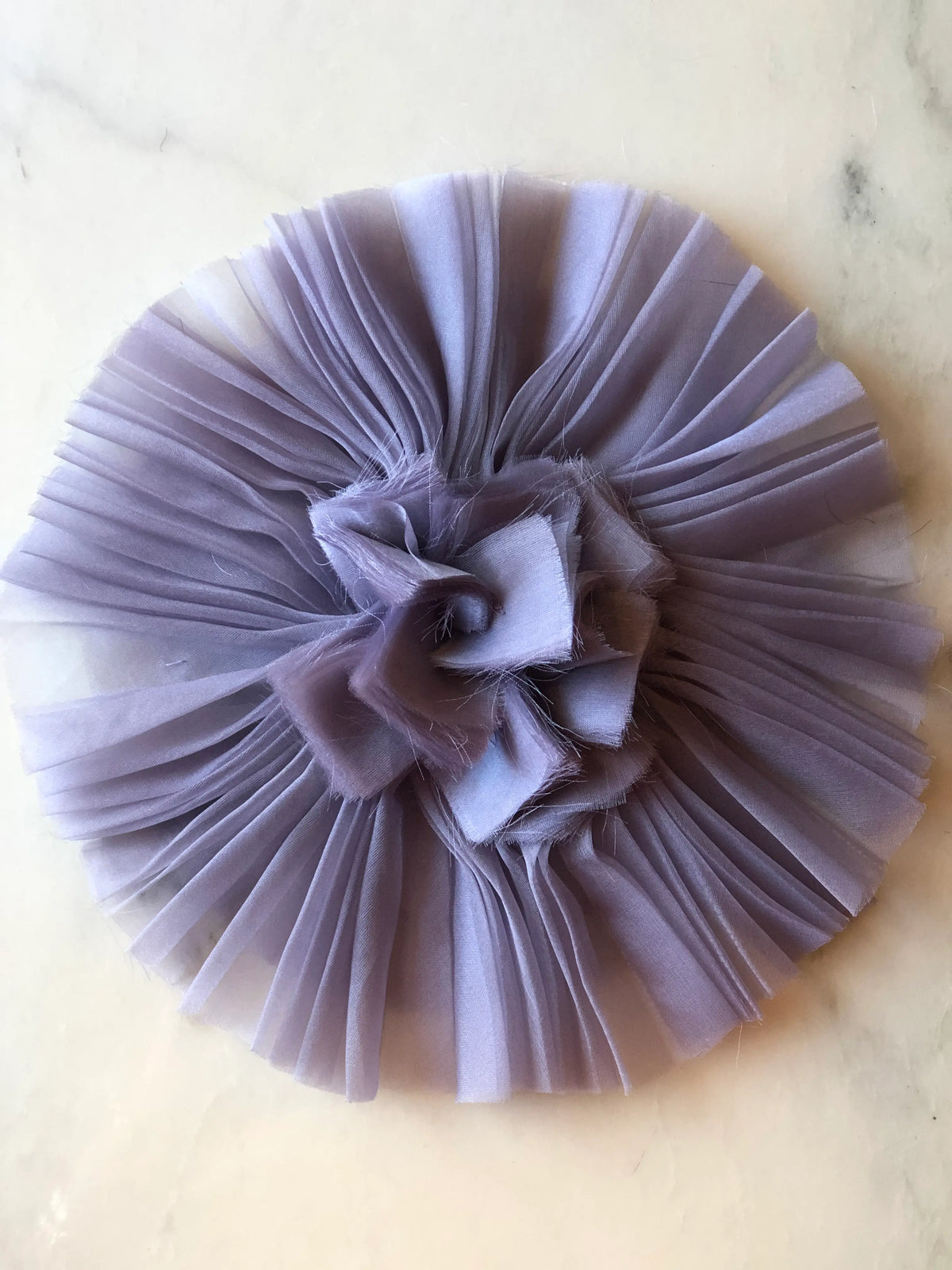Broochella Pieghettato Large Organza Flower Brooch Pin in soft lavender, featuring pleated organza petals arranged in a circular design with a softly frayed sculptural centre on a marble background.