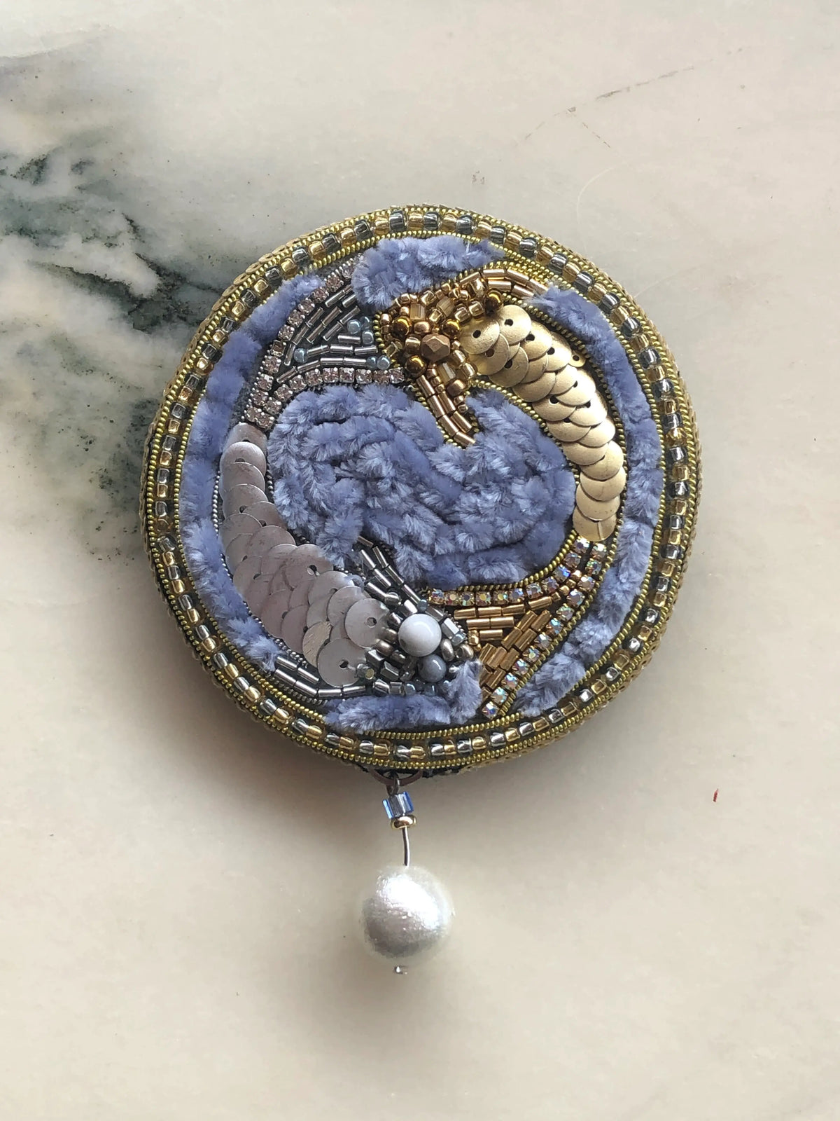 Broochella hand beaded Zodiac Pisces brooch pin featuring two beaded fish with a blue background and gold beads. The background is white marble