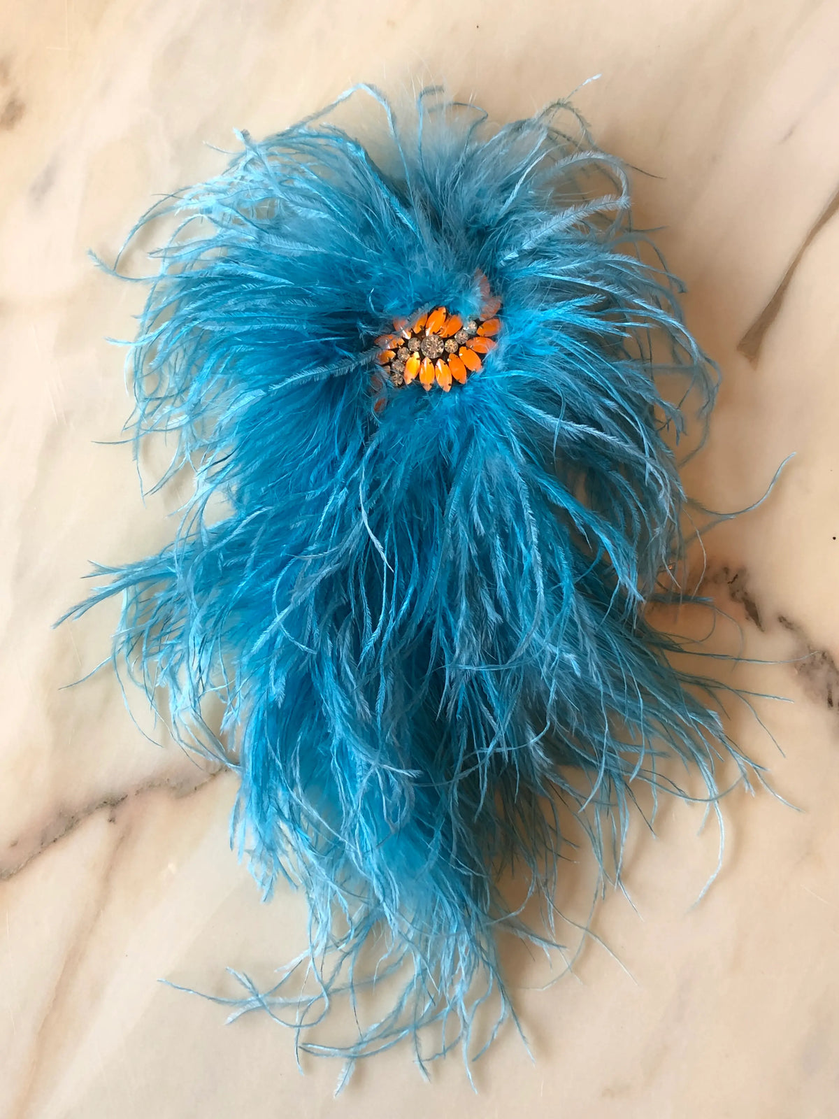 Broochella Piuma Neon Feather Brooch Pin in vibrant blue ostrich feathers with a contrasting orange jewelled centre, photographed on a white marble background.