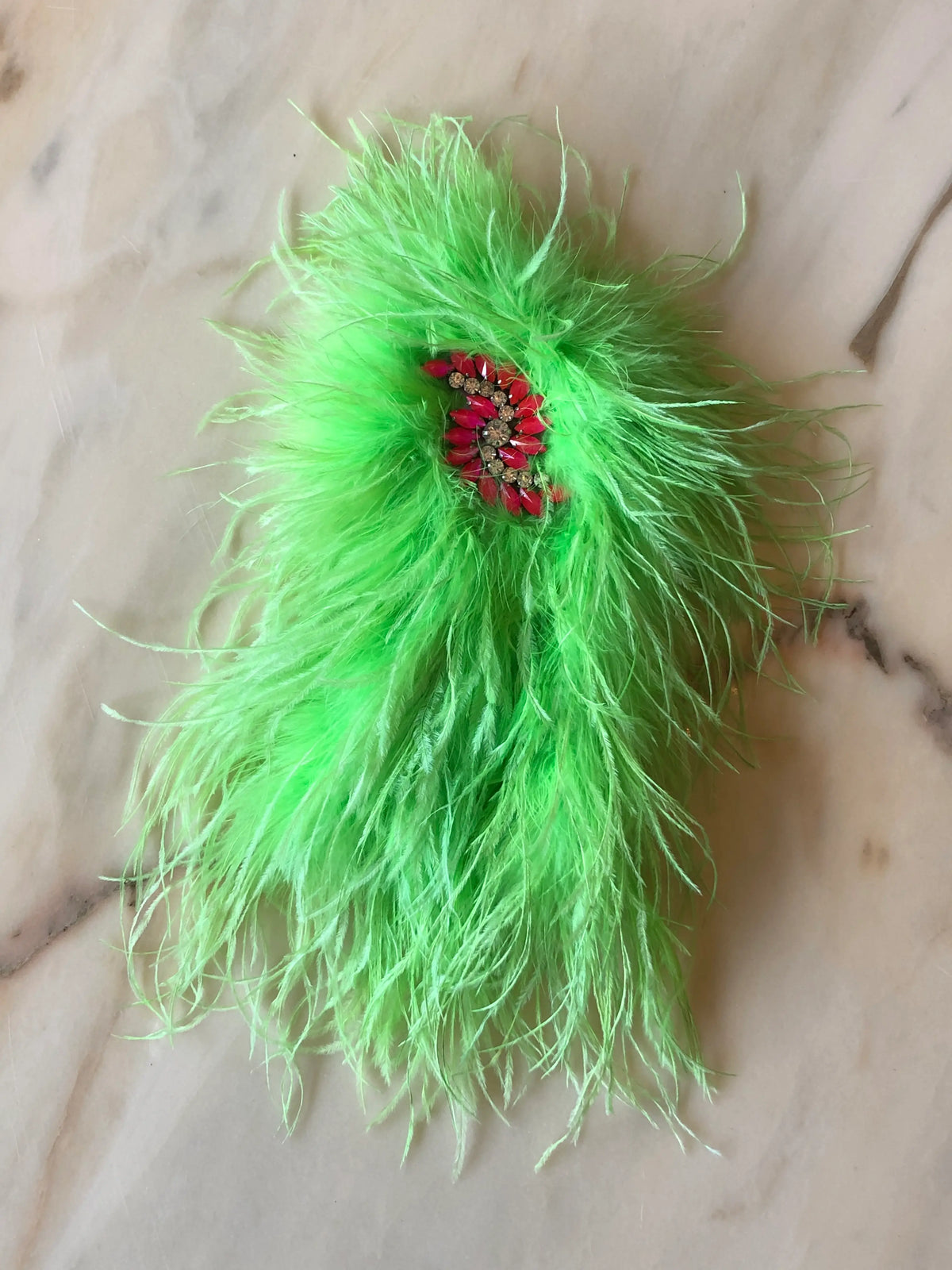 Broochella Piuma Neon Feather Brooch Pin in vibrant neon green ostrich feathers with a contrasting pink jewelled centre, photographed on a white marble background.