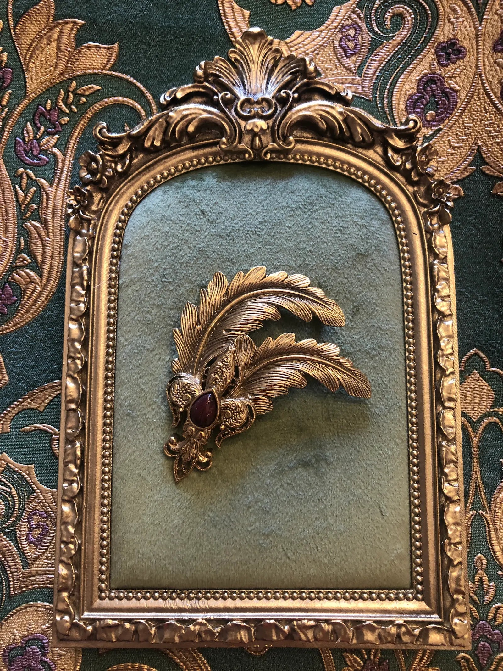 A Broochella vintage plume imperiale ornate gold-toned feather brooch with a dark red gemstone, displayed on a green textured panel within an elaborate gold frame, set against a patterned green and gold damask background.