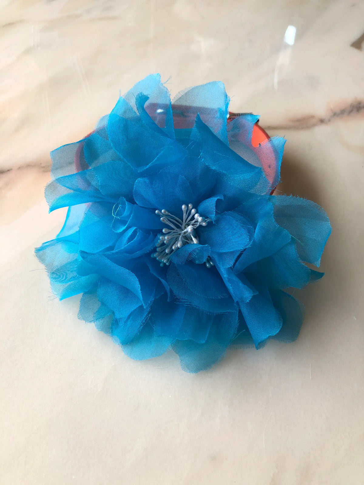 Broochella Poochella Aubrieta blue pet flower featuring layered turquoise sheer fabric petals and delicate white pearl-like stamens, attached to an orange dog collar with silver hardware, photographed on cream marble with natural grey veining.