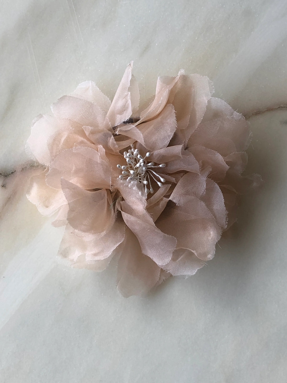 Broochella Poochella Aubrieta peach pet flower featuring airy, layered blush-peach sheer petals and white pearl-like stamens, photographed from above on a cream marble background with subtle grey veining.