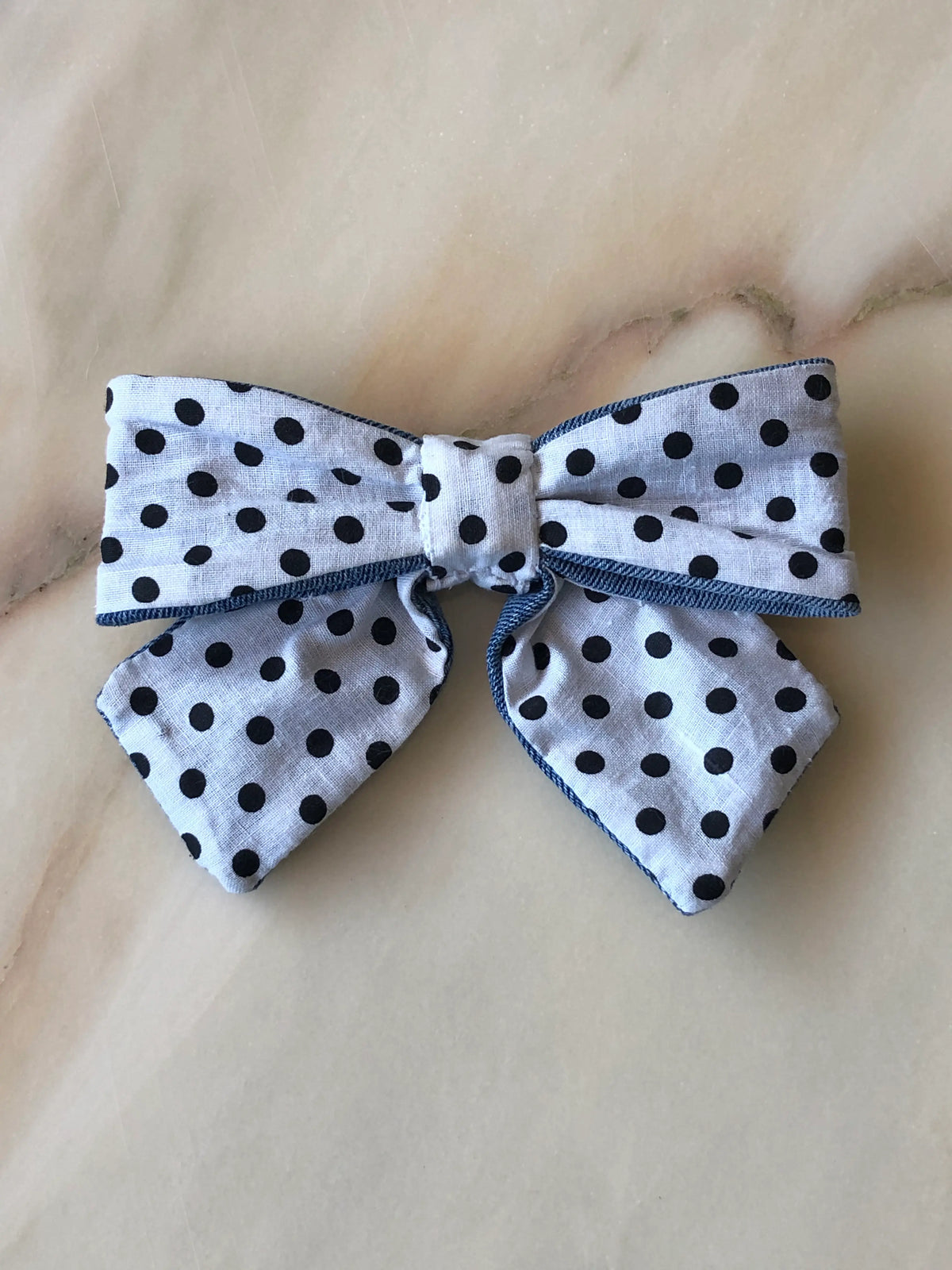 Broochella Poochella black polka dot dog bow in white cotton, featuring oversized loops, pointed tails and blue denim edging, photographed on a polished cream marble background with subtle beige and grey veining.