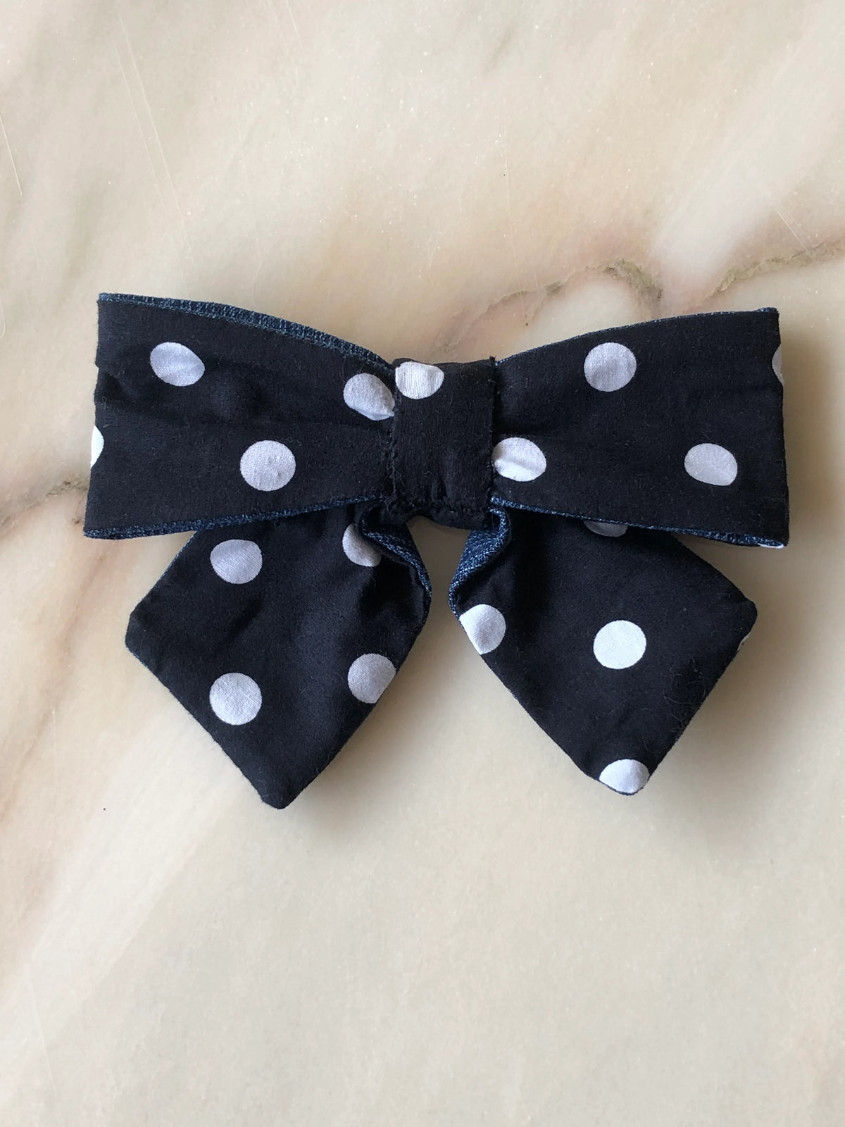 Broochella Poochella black and white polka dot pet bow featuring soft cotton fabric, contrasting blue denim trim, structured loops, a gathered centre and two pointed tails, photographed flat on polished cream marble with delicate beige and grey veining.