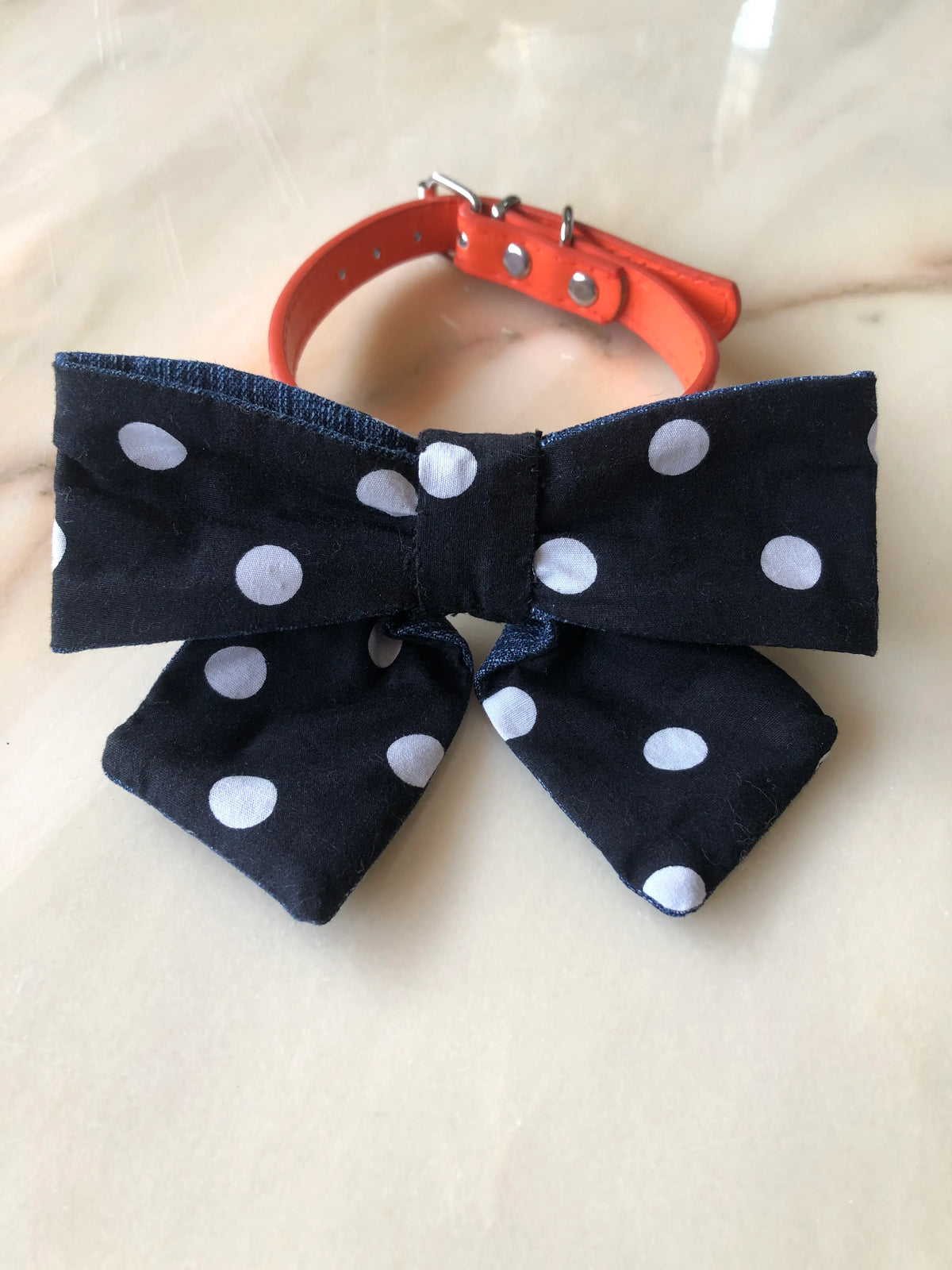 Broochella Poochella black cotton dog bow with large white polka dots, contrasting blue denim edging, a gathered centre and pointed tails, attached to an orange pet collar with silver hardware, photographed on polished cream marble with subtle beige and grey veining.