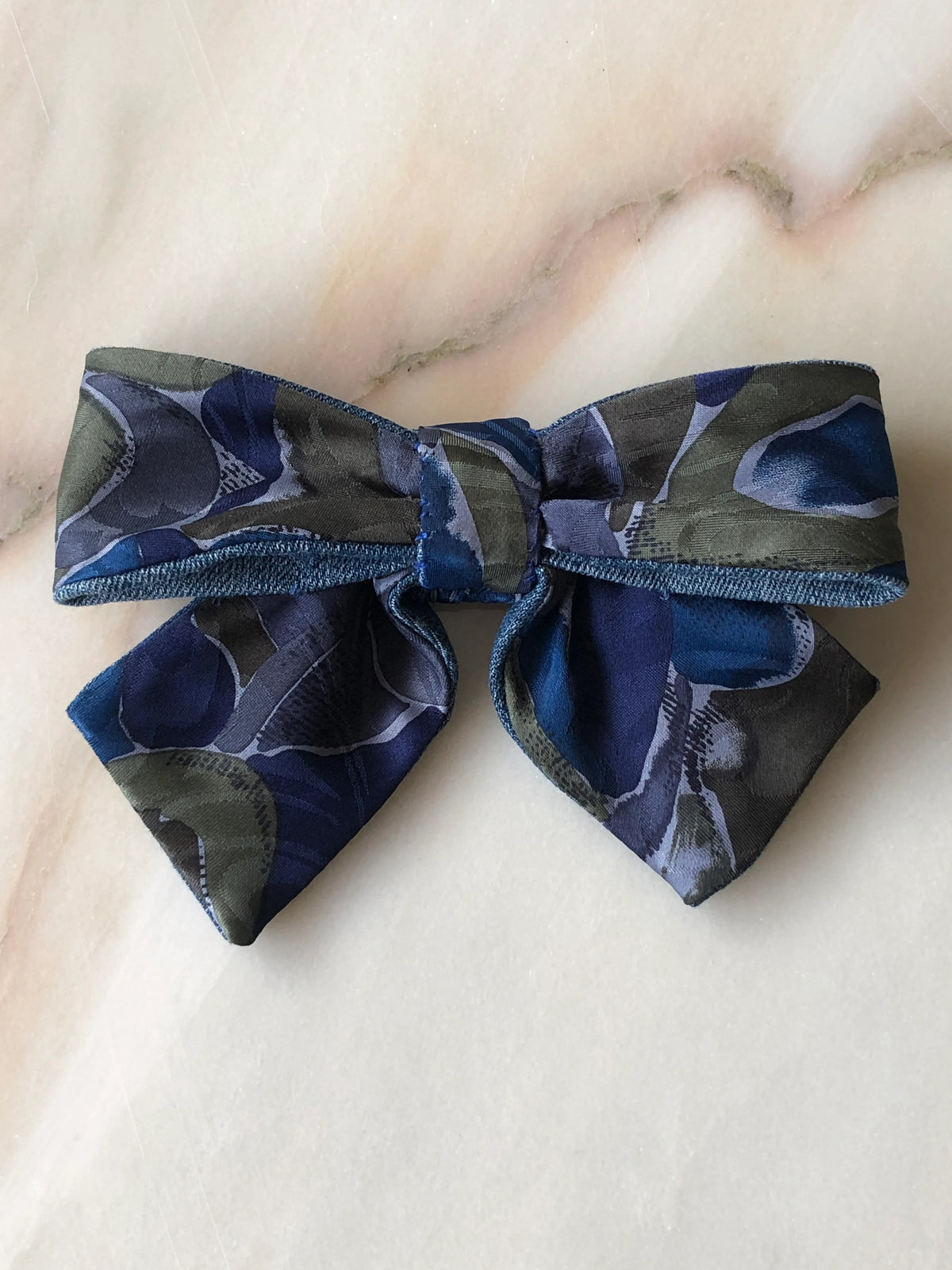 Broochella Poochella blue and green print pet bow featuring a bold abstract botanical pattern in deep navy, cobalt blue, olive green and muted grey, with a gathered centre, contrasting denim trim and two pointed tails, photographed flat on polished cream marble with delicate beige and grey veining.