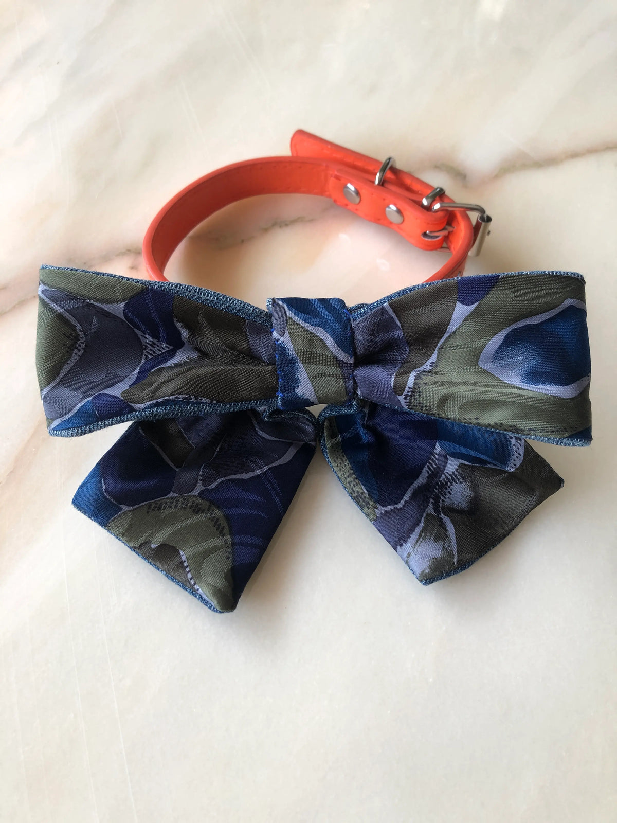 Broochella Poochella blue and green print dog bow featuring an abstract floral pattern in navy, cobalt blue, olive green and slate grey, finished with contrasting blue denim edging and pointed tails, attached to an orange pet collar with silver hardware, photographed on polished cream marble with subtle beige and grey veining.