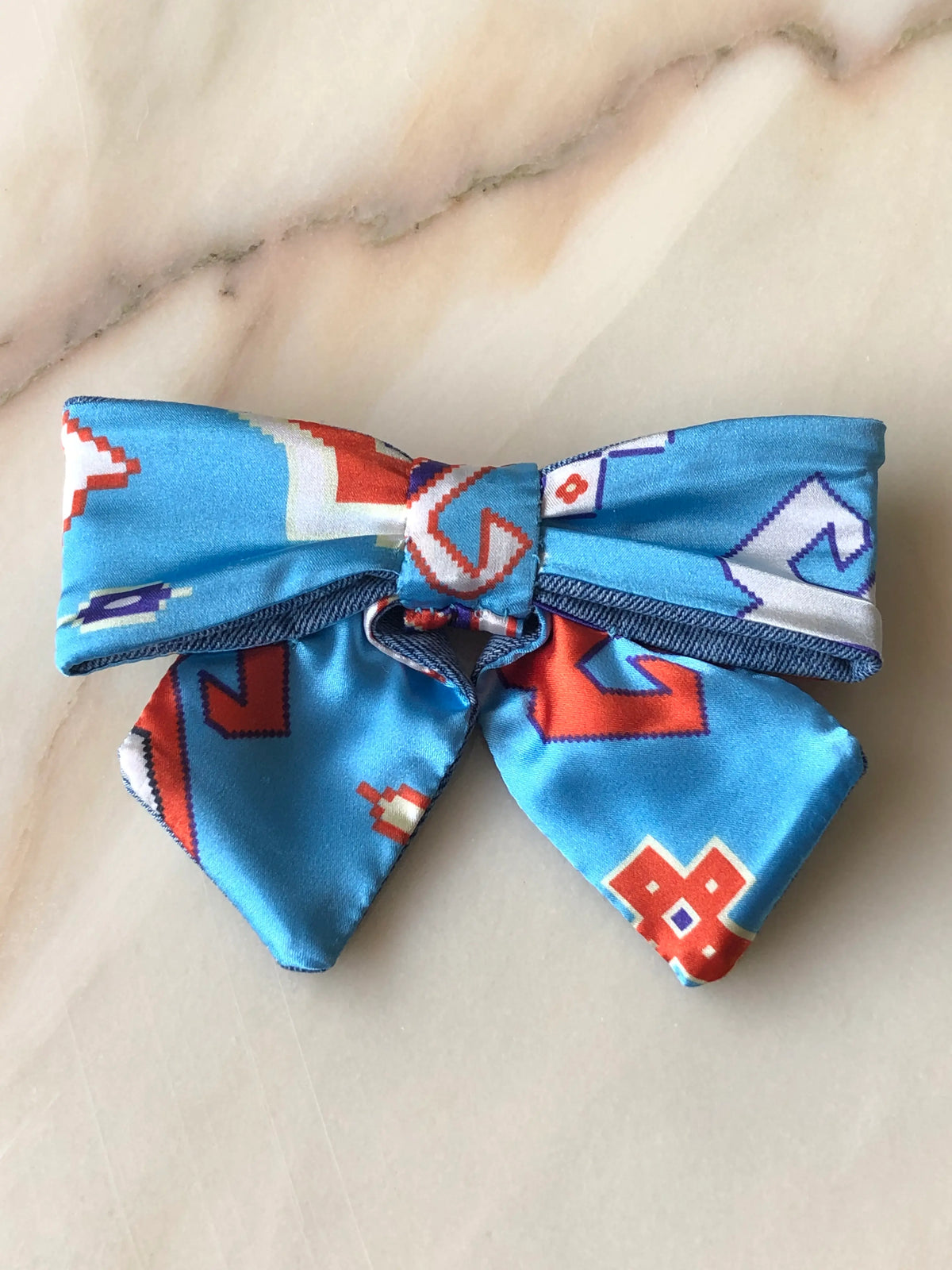 Broochella Poochella blue satin pet bow with a colourful abstract geometric print in orange, white and purple, contrasting denim trim, structured loops, a gathered centre and two pointed tails, photographed flat on polished cream marble with delicate beige and grey veining.