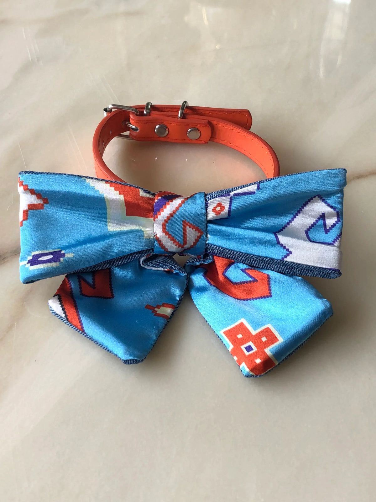 Broochella Poochella blue satin pet bow with a colourful abstract geometric print in orange, white and purple, contrasting denim trim, structured loops, a gathered centre and two pointed tails, photographed flat on polished cream marble with delicate beige and grey veining.