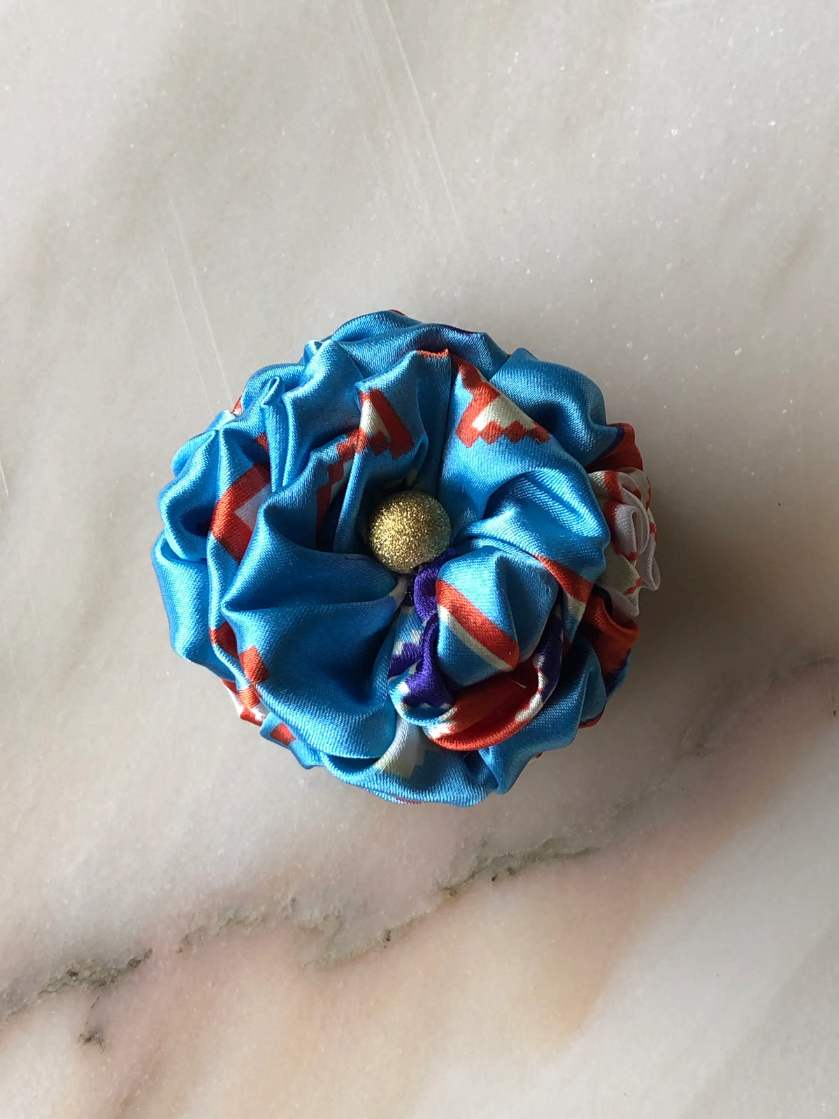Broochella Poochella Bougainville Positano blue satin pet flower featuring gathered petals with orange, white and purple abstract patterns and a textured gold centre, photographed from above on a polished cream marble background with subtle grey veining.