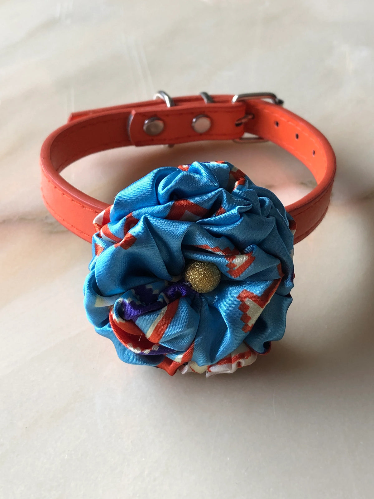 Broochella Poochella Bougainville Capri pet flower collar featuring a gathered satin flower with pink, orange, cream and black abstract print, a textured gold centre and an adjustable orange collar with silver hardware, photographed on a polished cream marble background with subtle grey veining.