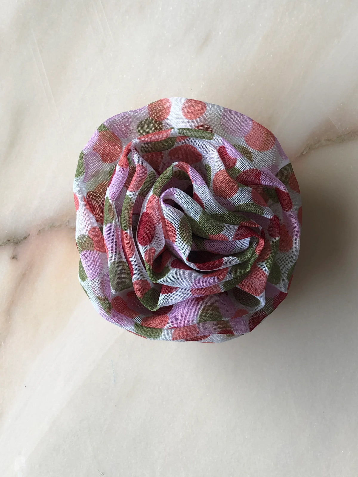 Broochella Poochella Brassica pink pet flower featuring a voluminous rosette of softly gathered sheer white mesh with pink, coral, lilac and olive-green polka dots, photographed from above on cream marble with subtle grey veining.