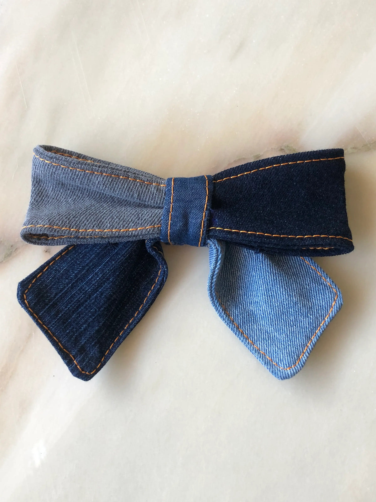 Broochella Poochella denim pet bow featuring contrasting shades of light blue, charcoal grey and dark indigo denim, a gathered centre, structured loops, pointed tails and decorative orange stitching, displayed on polished cream marble with soft beige and grey veining.