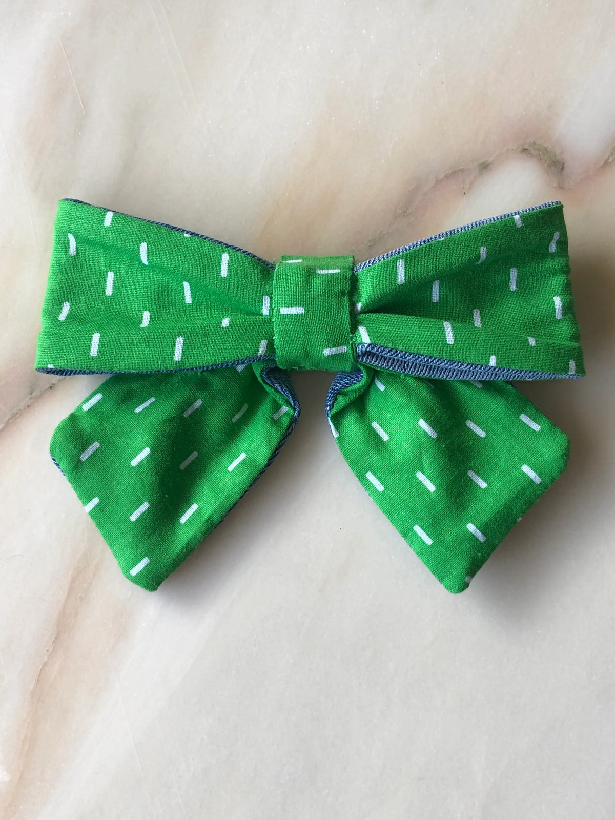 Broochella Poochella bright green cotton pet bow with scattered white dash print, structured loops, gathered centre, contrasting blue denim trim and two pointed tails, photographed flat on polished cream marble with delicate beige and grey veining.