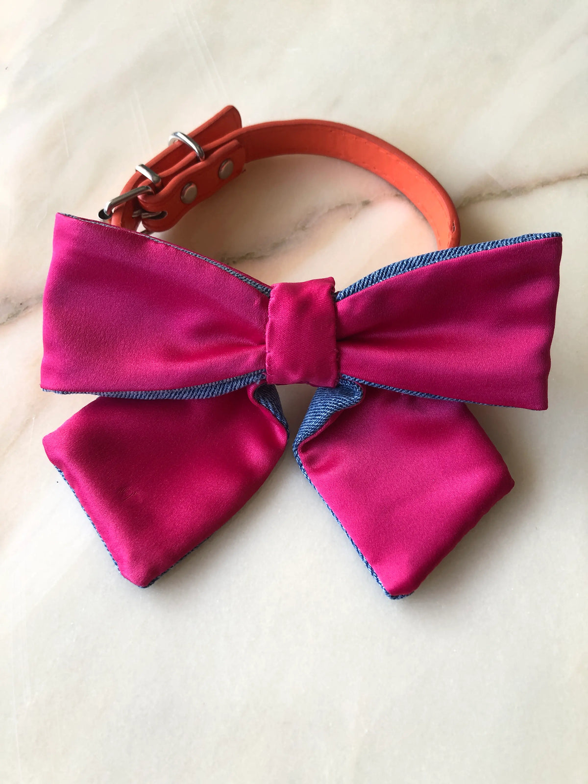 Broochella Poochella pink satin dog bow featuring vibrant fuchsia satin, contrasting blue denim edging, a gathered centre and pointed tails, attached to an orange pet collar with silver hardware, photographed on polished cream marble with subtle beige and grey veining.