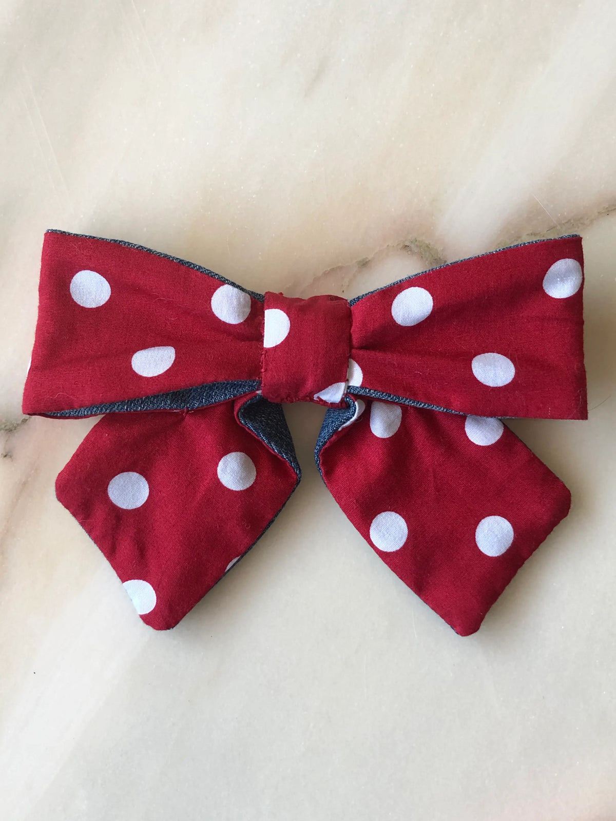 Broochella Poochella red and white polka dot cotton dog bow featuring oversized loops, a gathered centre, pointed tails and contrasting blue denim edging, displayed on a polished cream marble background with soft beige and grey veining.