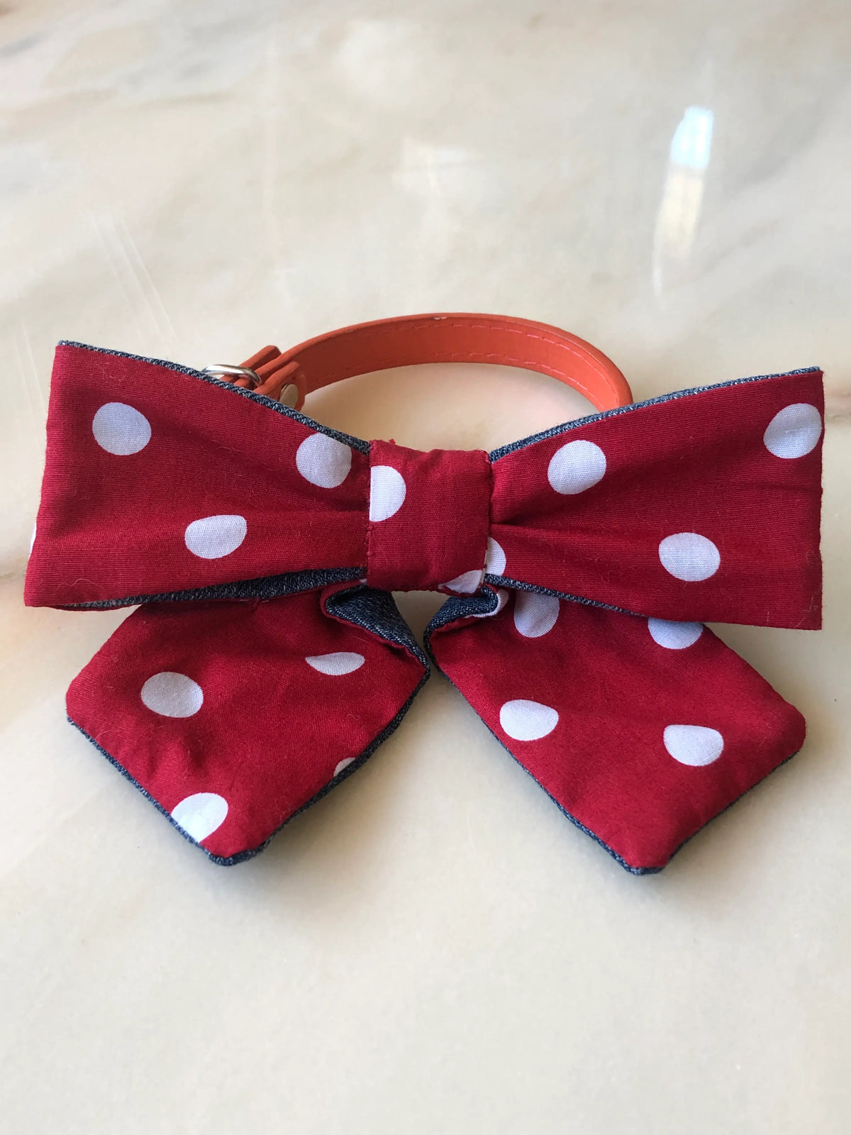 Broochella Poochella red cotton dog bow with large white polka dots, blue denim edging and pointed tails, attached to an orange pet collar with silver hardware, photographed on a polished cream marble background with subtle beige and grey veining.
WebP Filename: