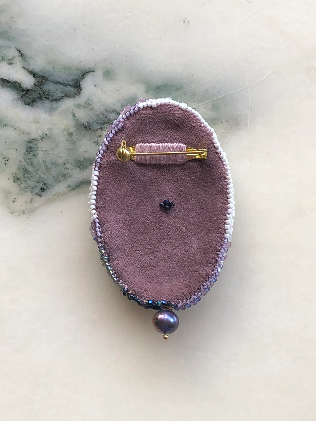 The back view of Broochella Portrait of Aphrodite Beaded Brooch featuring an intricately hand-beaded classical female figure in soft blush, ivory, silver, blue and bronze tones, photographed on a white marble background.